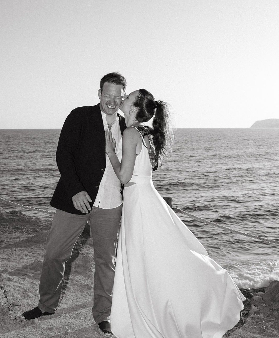 A couple dressed in wedding attire sharing a joyful moment by the ocean, with the woman kissing the man on the cheek.