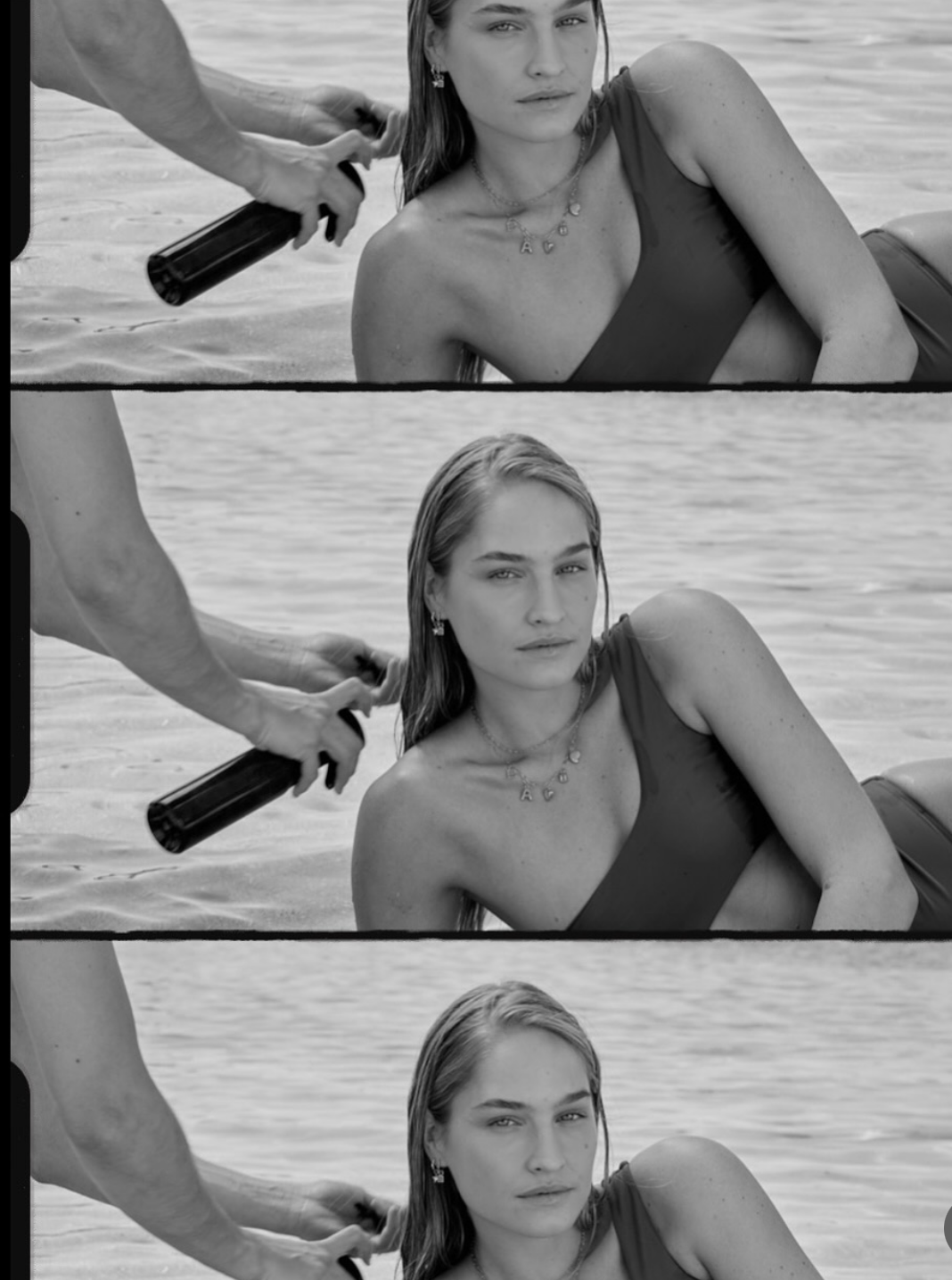 Black and white photo of a woman lying on a beach, with someone spraying her hair with a bottle of water, and ocean in the background.