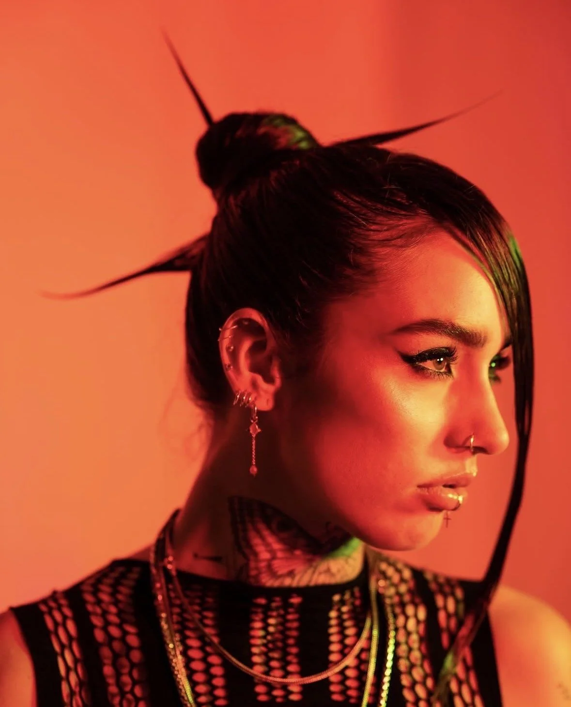 A woman with dark hair styled in a top knot, wearing multiple earrings, a nose ring, and layered necklaces, looking to the right against an orange-red background.