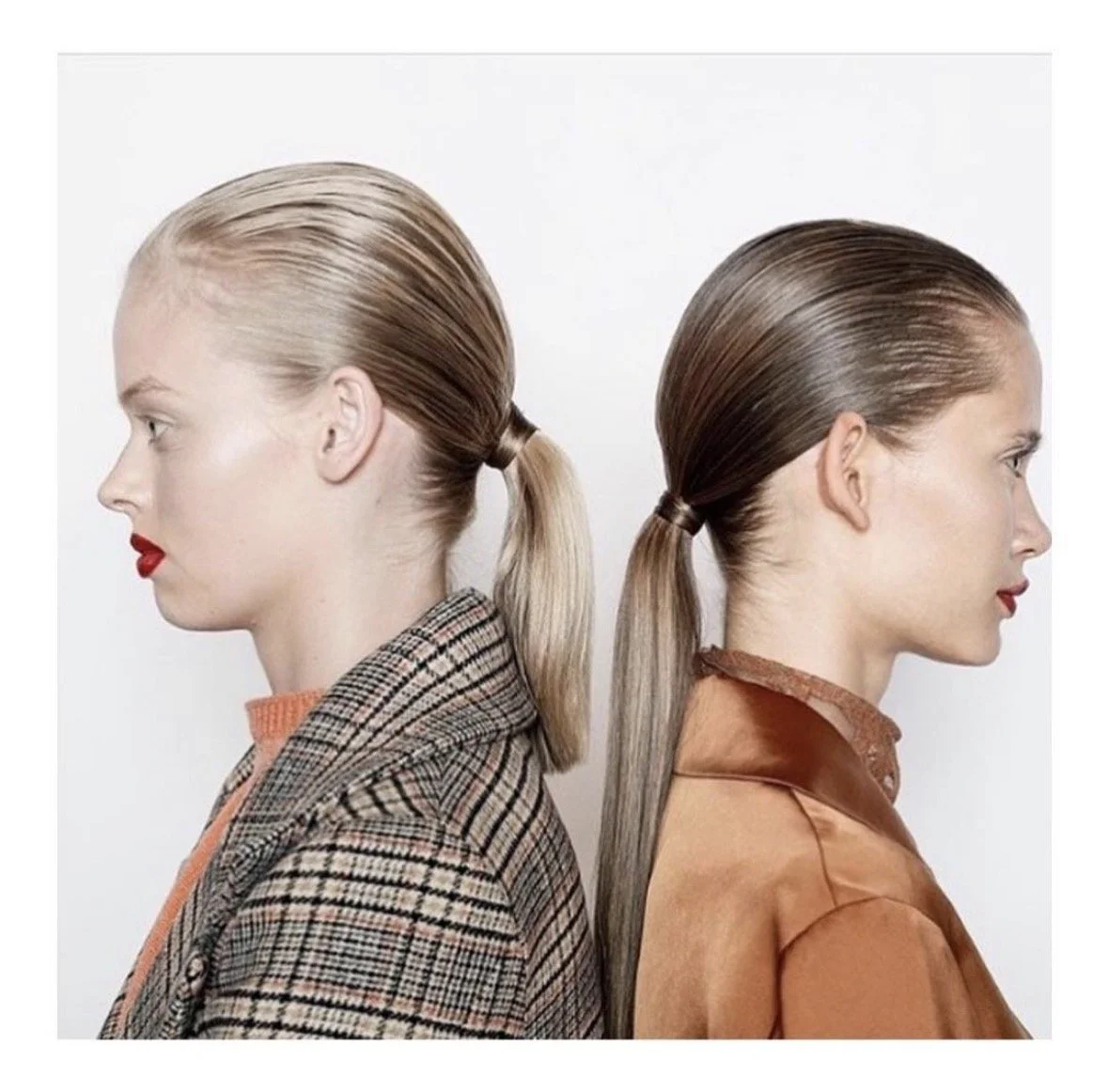 Side profile of two women with slicked-back hair tied in low ponytails, one with blonde hair and the other with brown hair, wearing stylish coats and red lipstick.