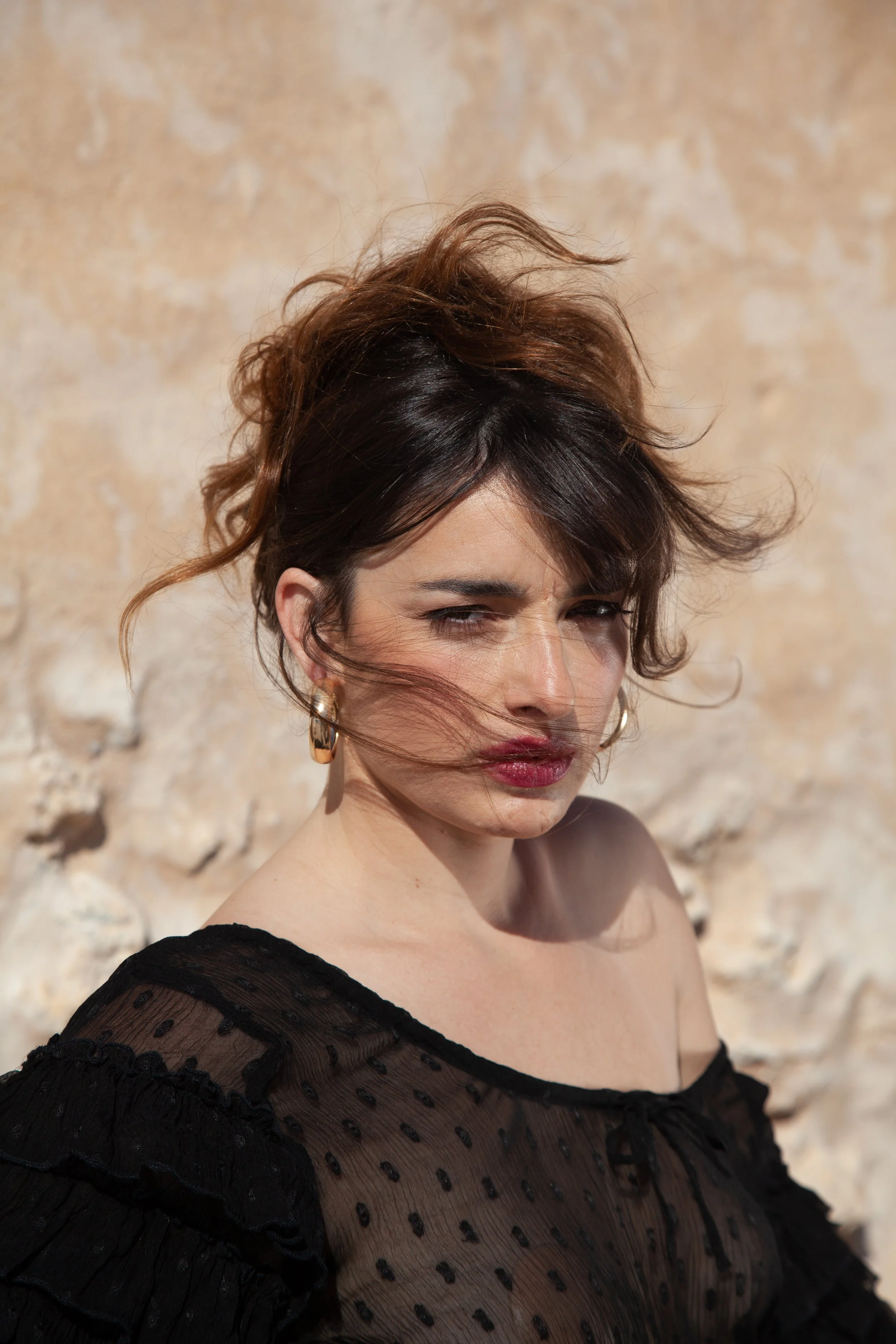 A woman with tousled brunette hair and makeup, wearing gold hoop earrings and a black sheer top with polka dots, standing in front of a textured beige wall.