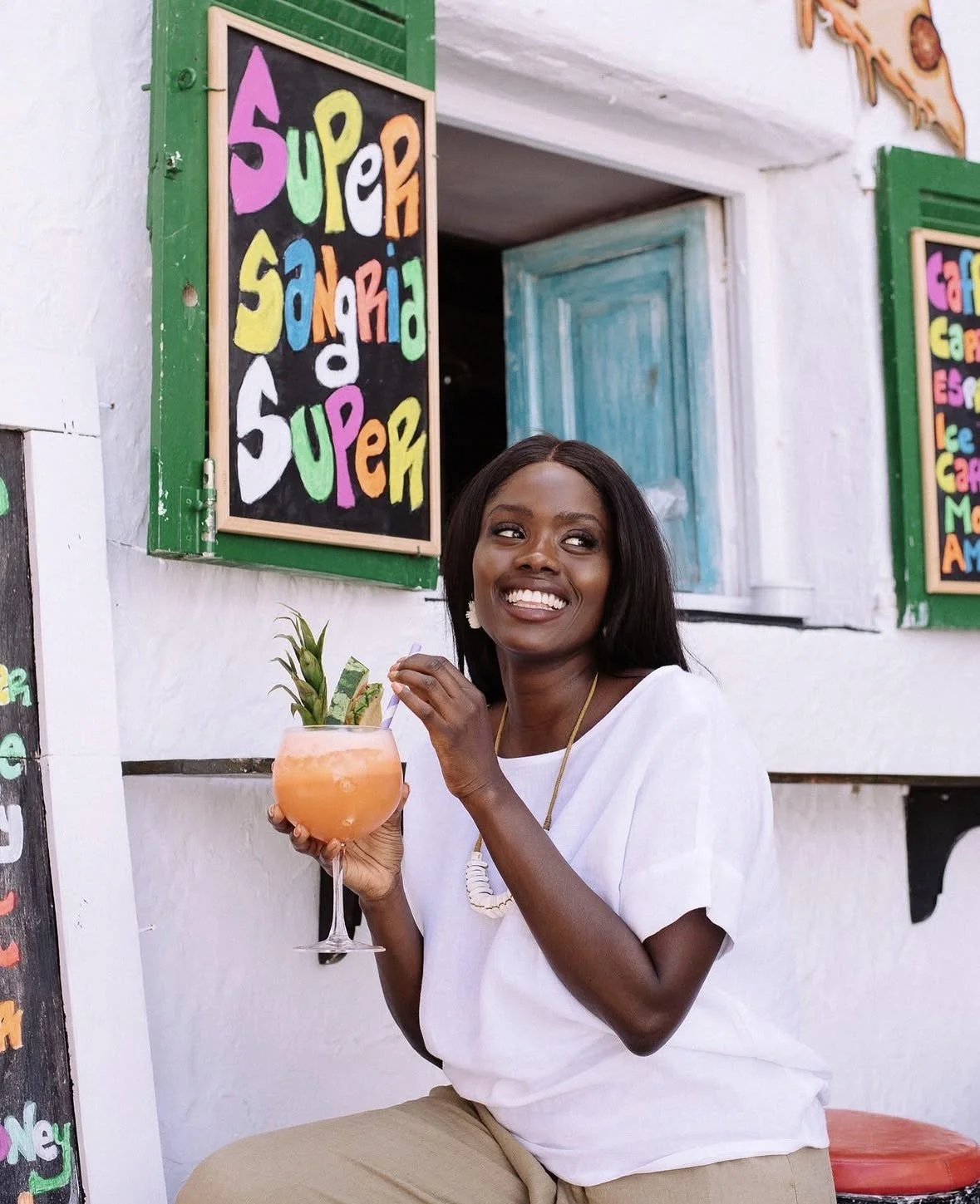 A woman with dark hair and a white top smiling and holding a pink cocktail garnished with a pineapple leaf outside a colorful restaurant with vibrant signs.