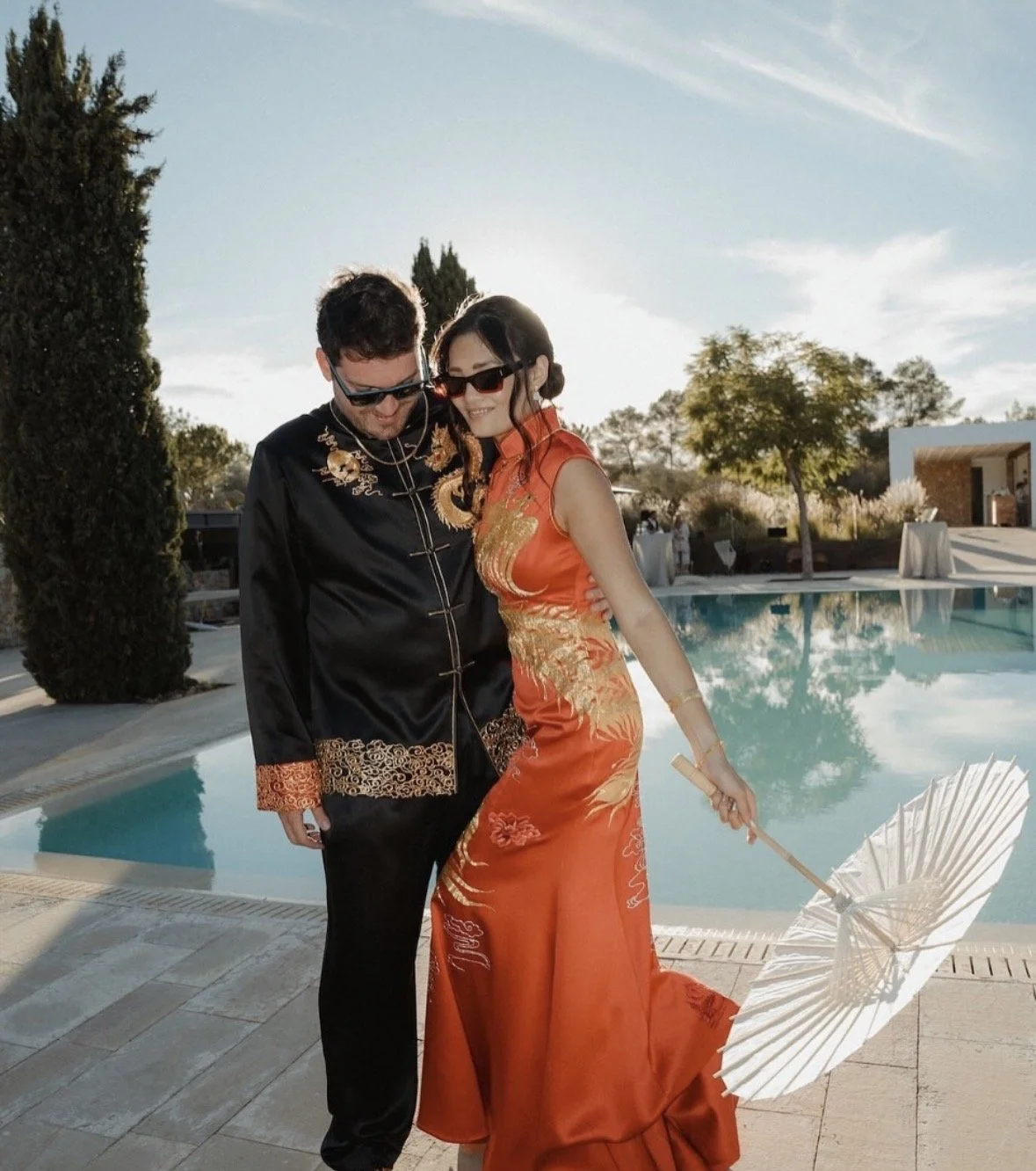 Couple dressed in traditional Asian attire standing by a pool, woman holding a white parasol, both wearing sunglasses and smiling in an outdoor setting.