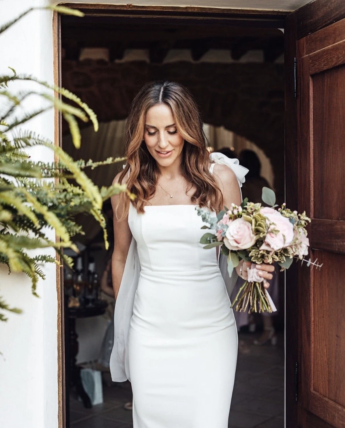 A bride in a white wedding dress holding a bouquet of pink and white flowers, standing at a doorway.