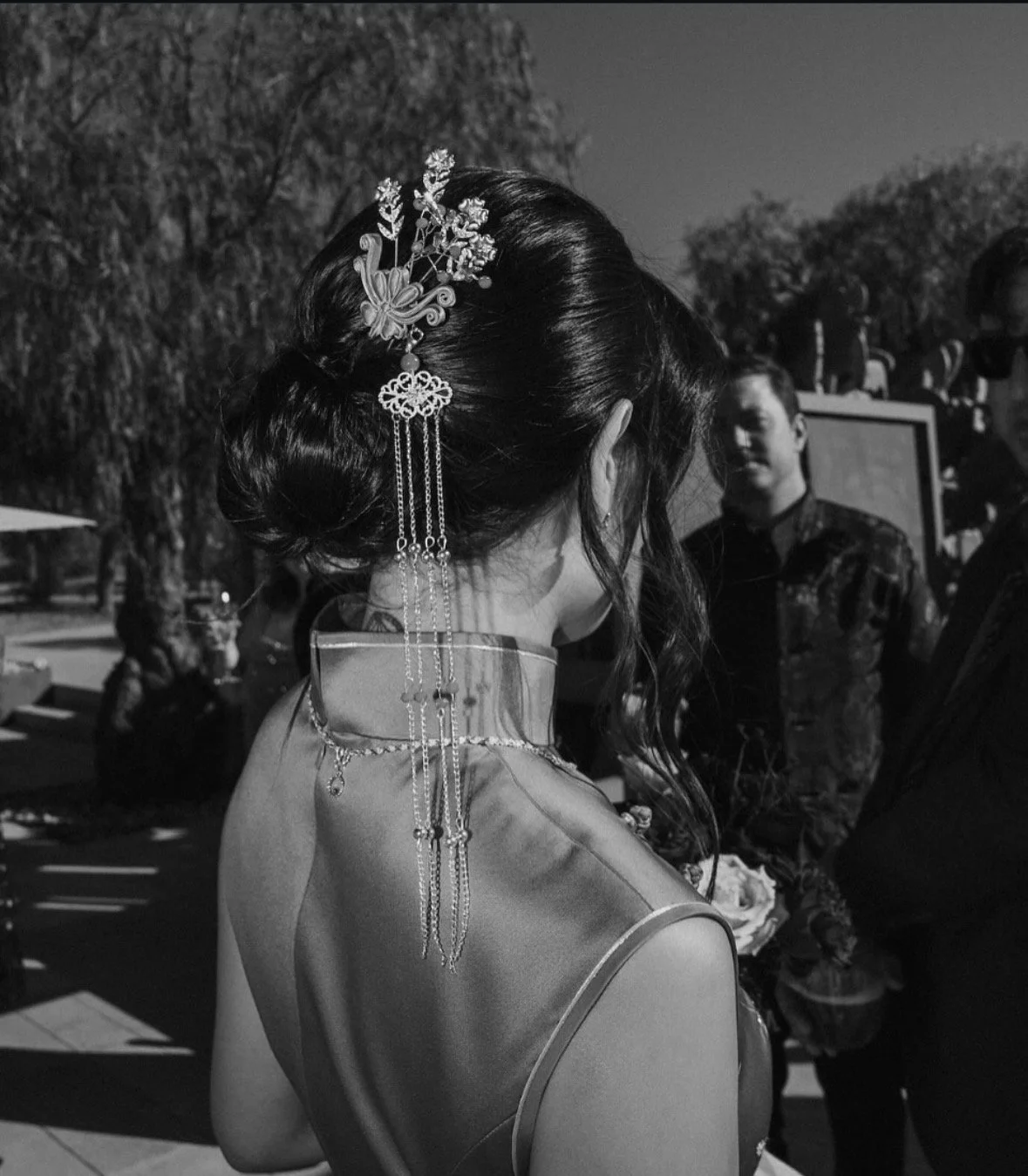 A woman with traditional hairstyle and hair accessories, wearing a garment made of shiny fabric, stands outdoors talking to a man. Several people are seen in the background.