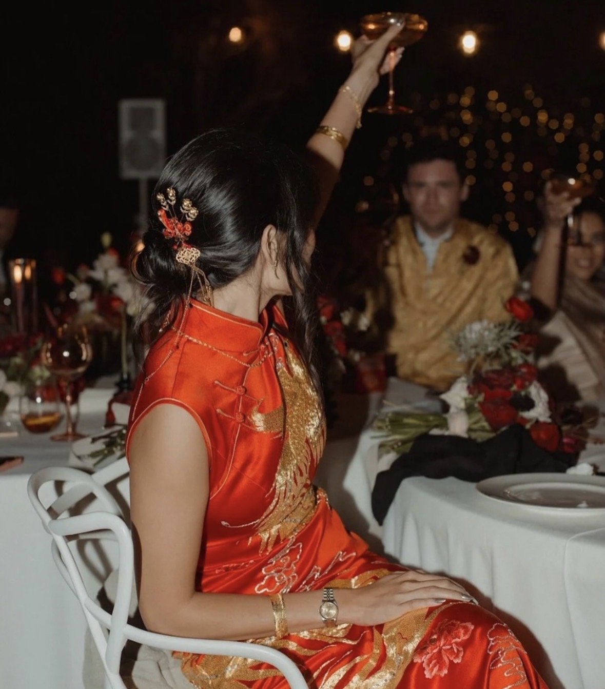 A woman in a traditional red and gold dress is celebrating at a dinner event, raising a glass in toast with other guests around her.