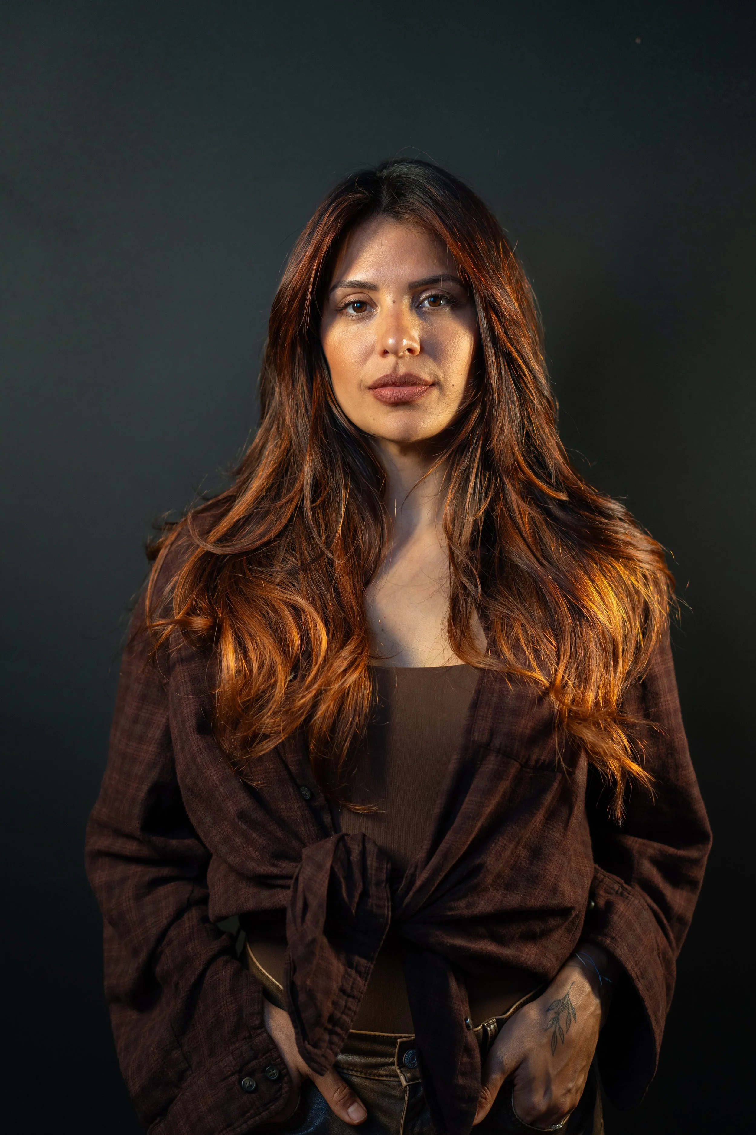 A woman with long, wavy brown hair standing against a dark background.