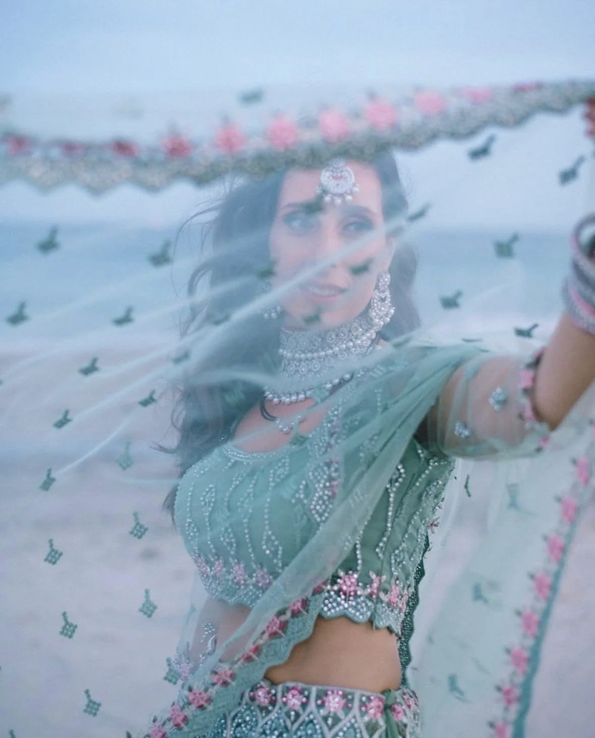 A woman in traditional Indian attire is looking through a sheer fabric with embroidery and pink accents, with her reflection visible. She is wearing jewelry and is outdoors.