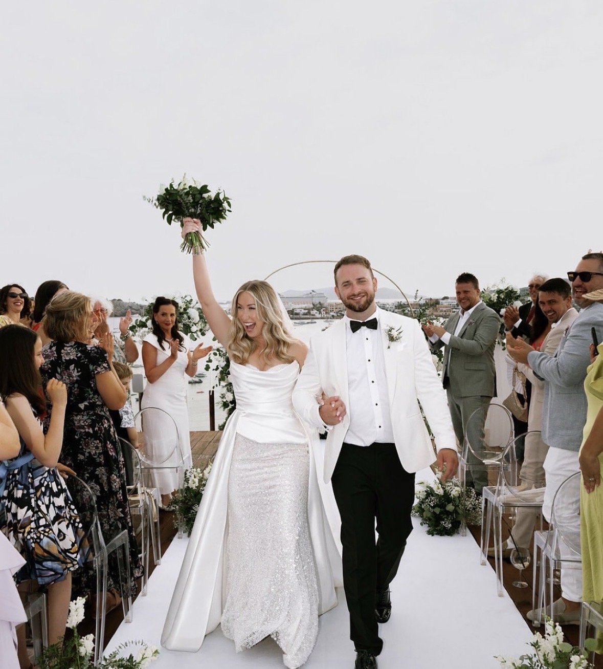 A newlywed couple walking down the aisle outdoors, celebrating their wedding, surrounded by smiling guests, with a waterfront in the background.
