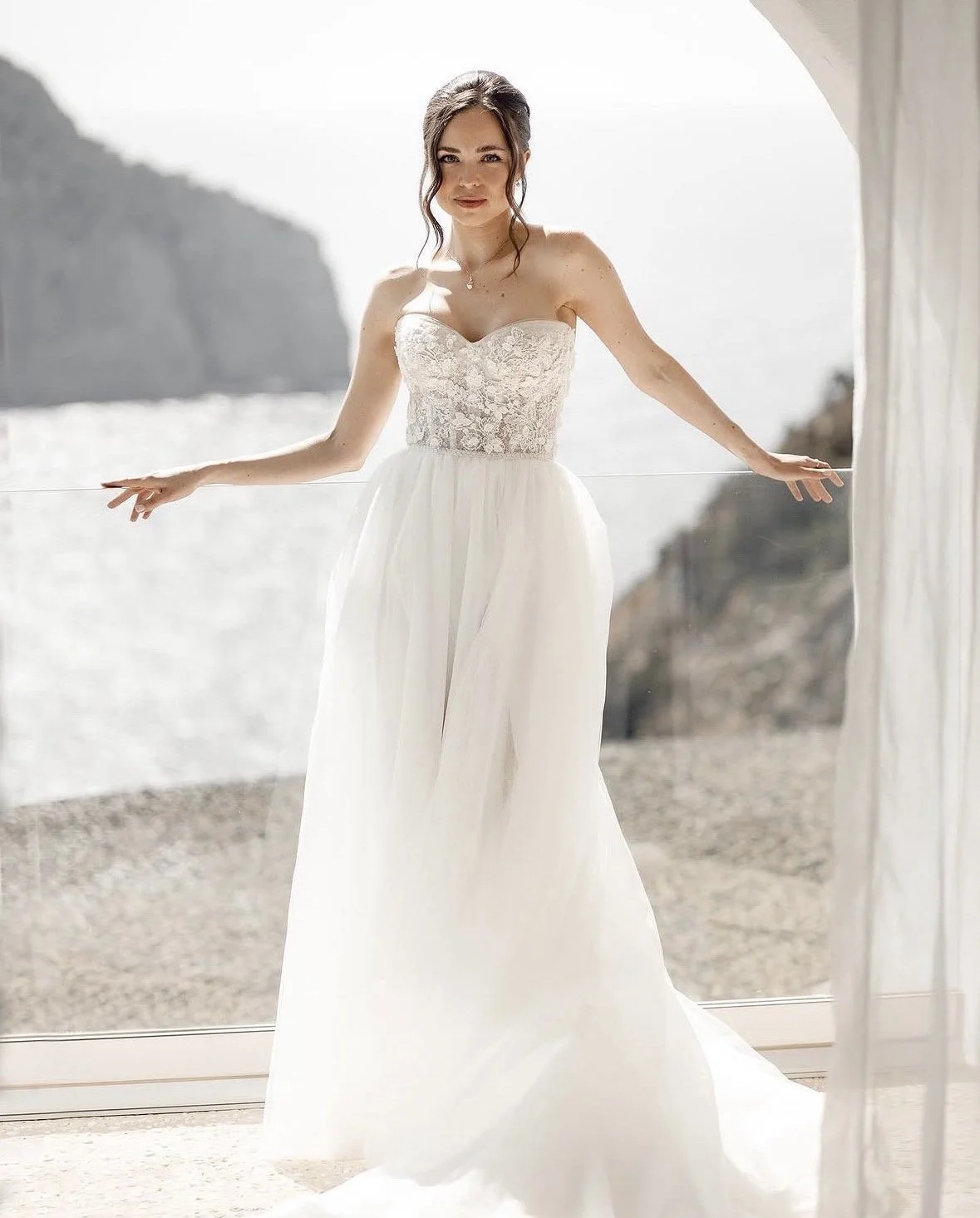 A woman in a wedding dress standing by a glass door overlooking the beach and rocky cliffs.