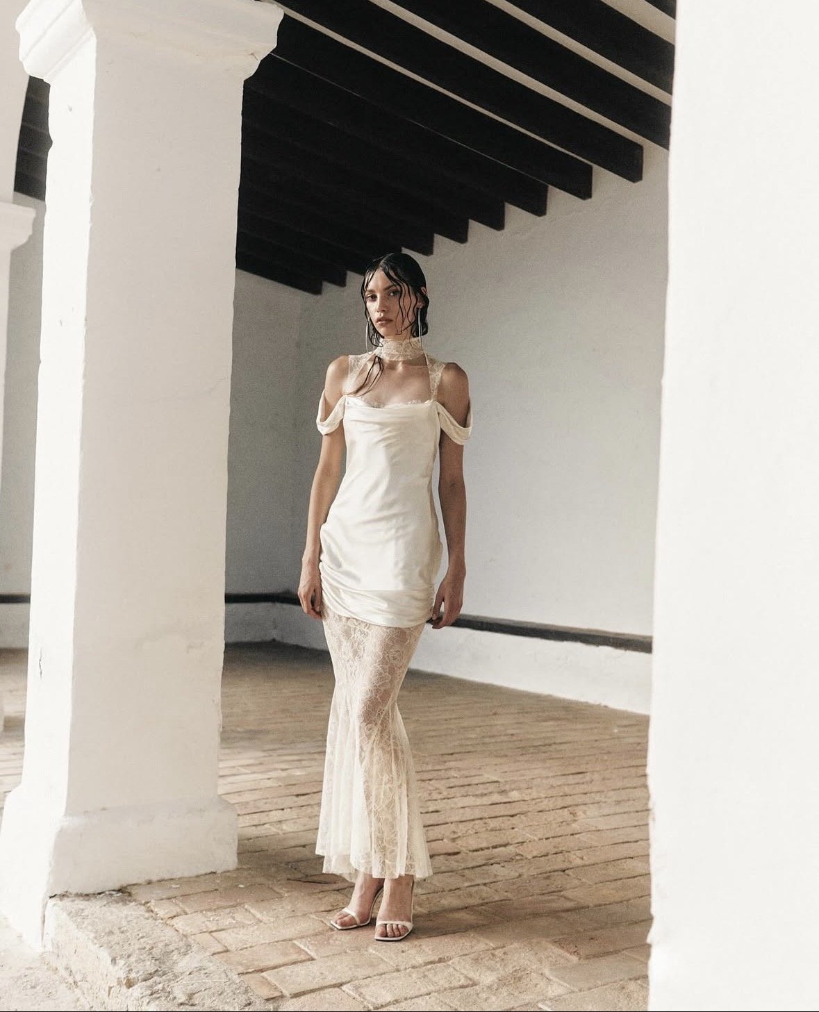 Woman in cream-colored dress with lace details standing indoors on brick floor under dark wooden beams.