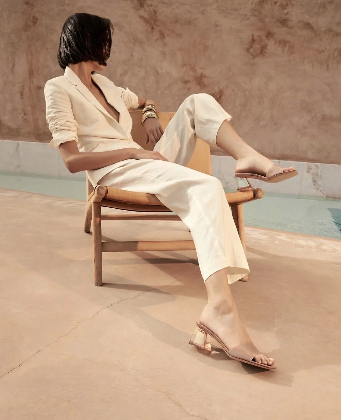 Woman sitting on a wooden chair beside a swimming pool, wearing a cream-colored suit and beige high-heeled sandals.