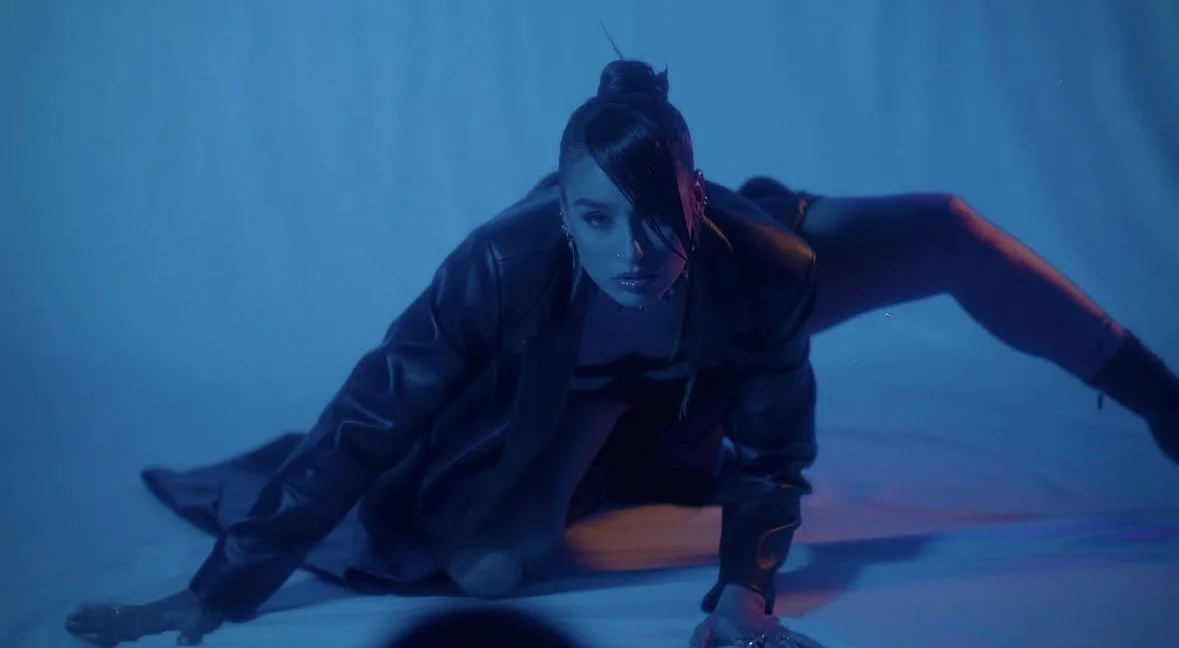 A woman with dark hair in a braid, dressed in black, is crawling on the ground in a dim, blue-lit setting.