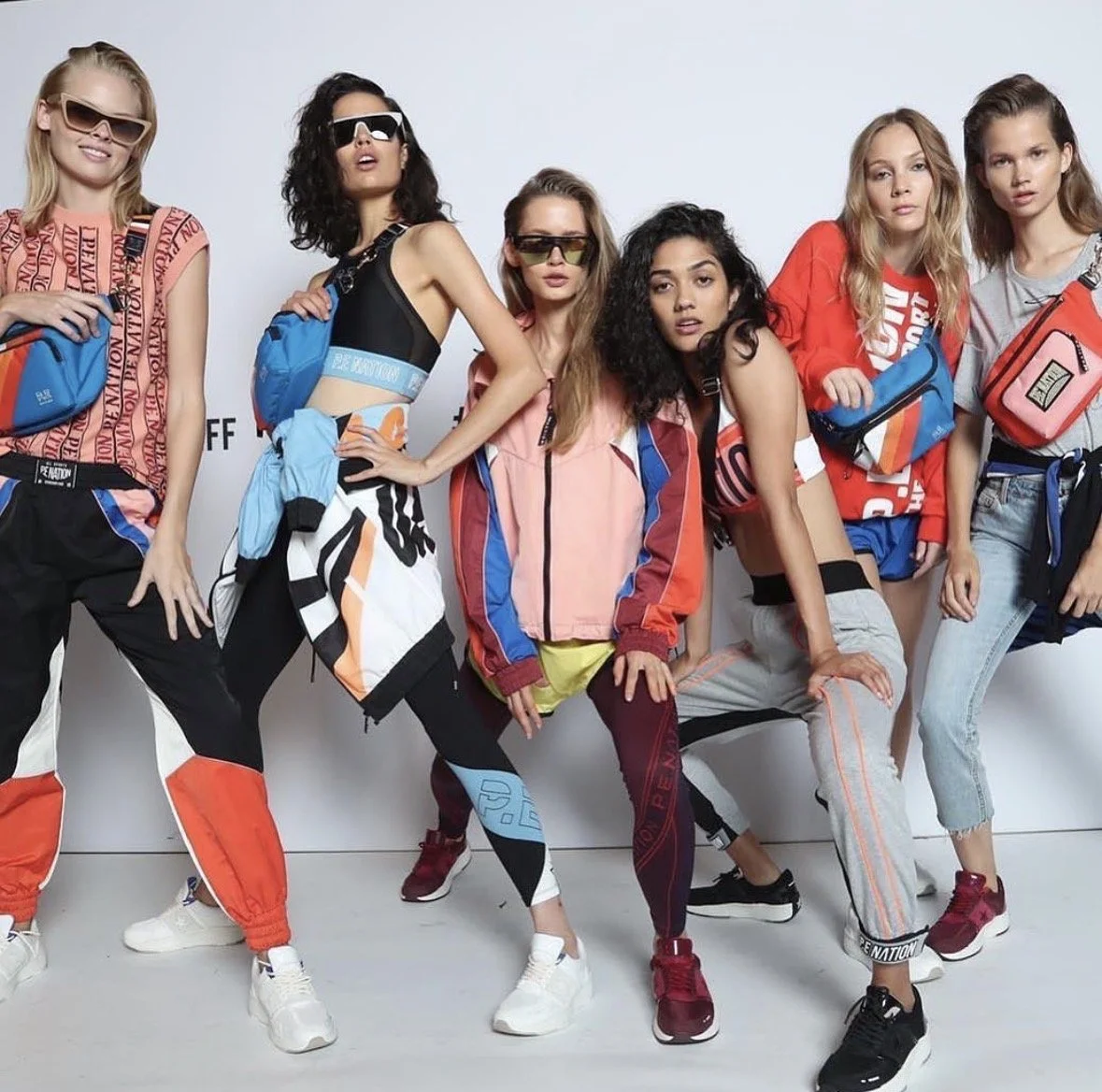 Group of young women in colorful sporty clothing and sunglasses, posing in front of a plain white backdrop, each carrying a small backpack.