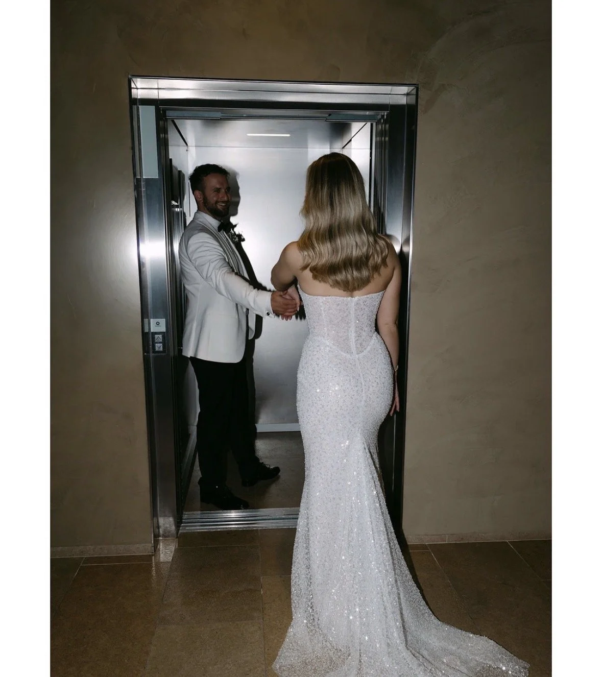 A bride and groom holding hands in an elevator, with the groom wearing a white tuxedo jacket and the bride in a strapless, sparkly wedding gown.