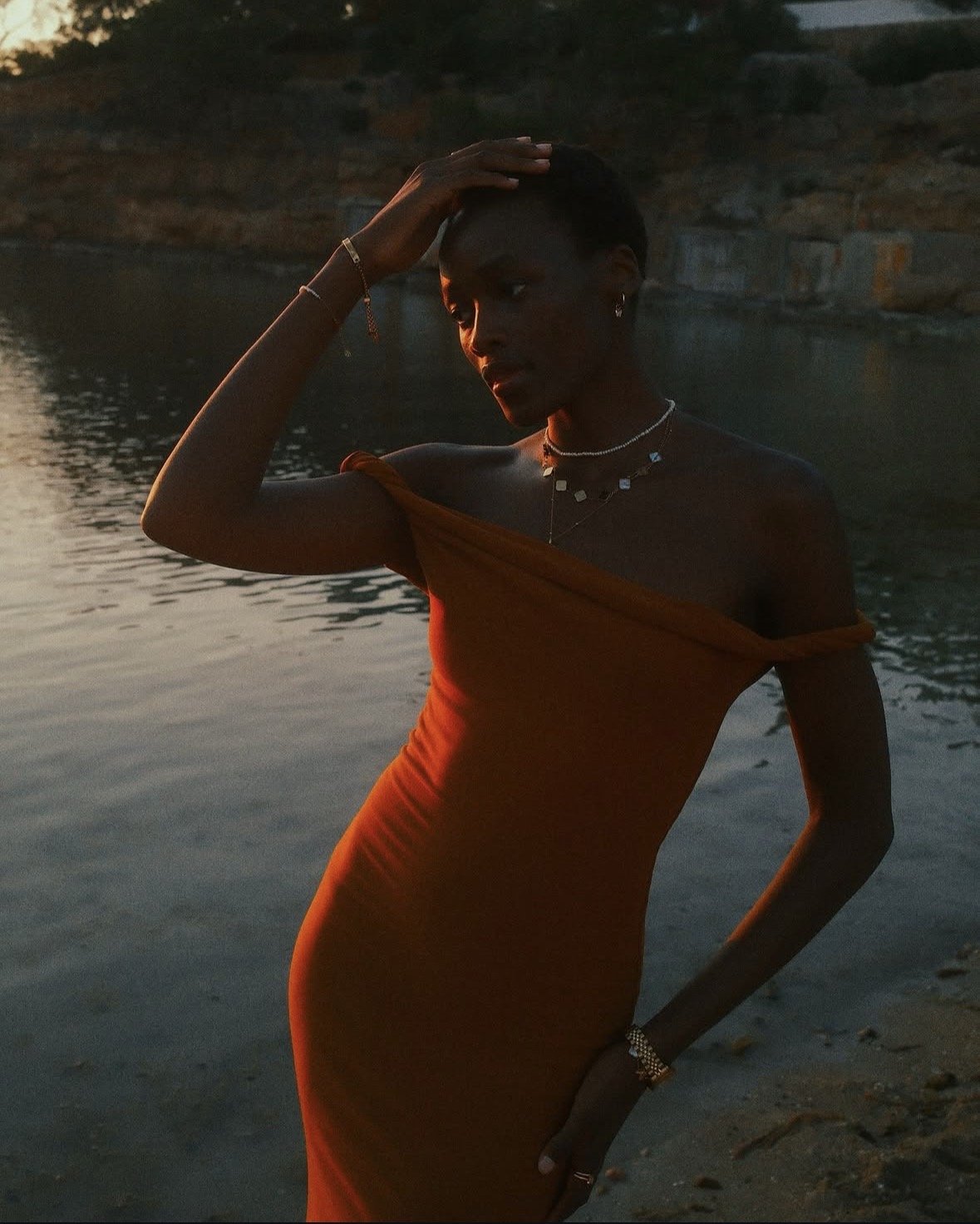 A woman in an orange dress standing near water during sunset, with her hand on her head and looking down.