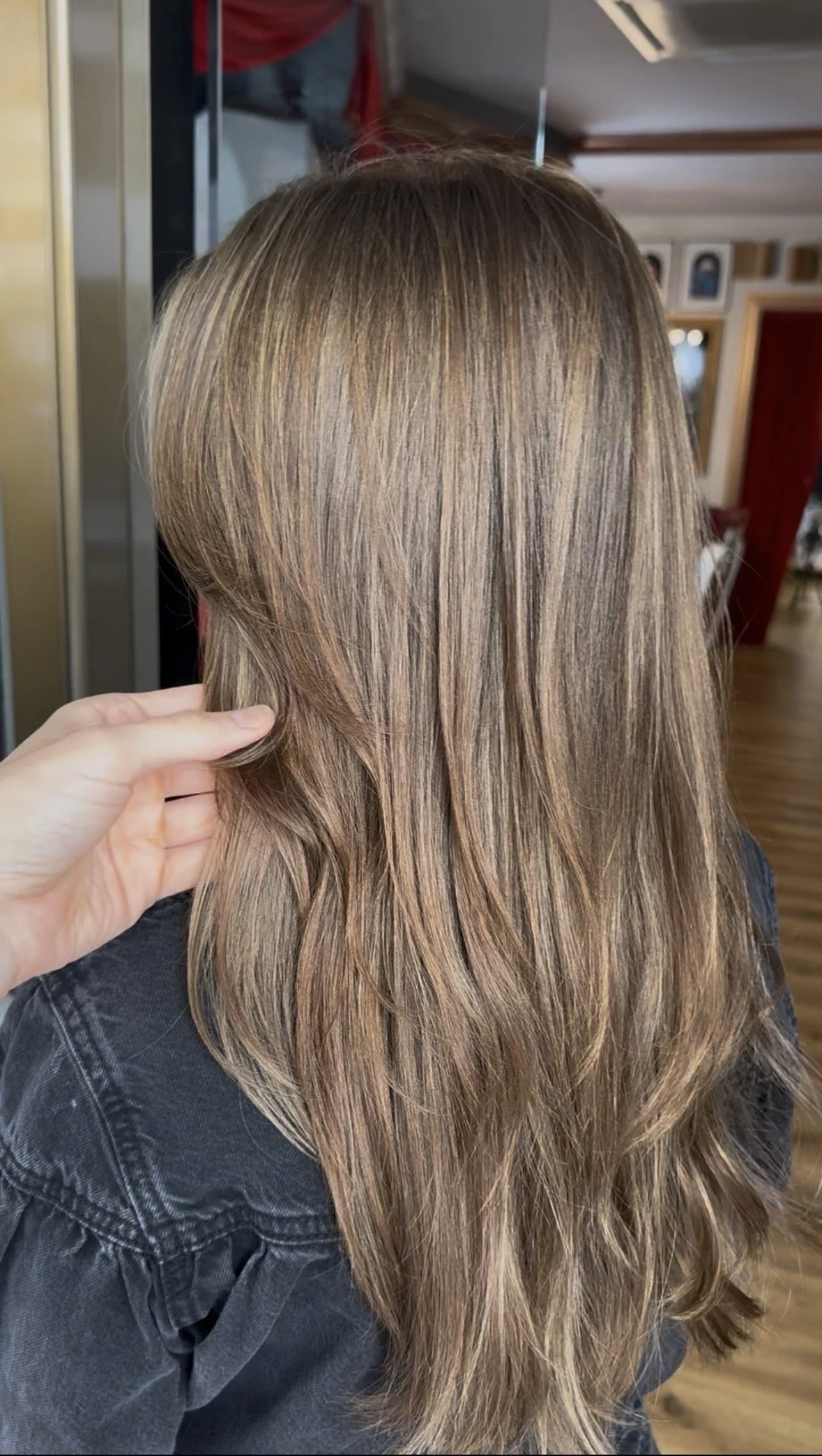 Back view of a woman with wavy, light brown hair; someone is touching her hair on the left side.