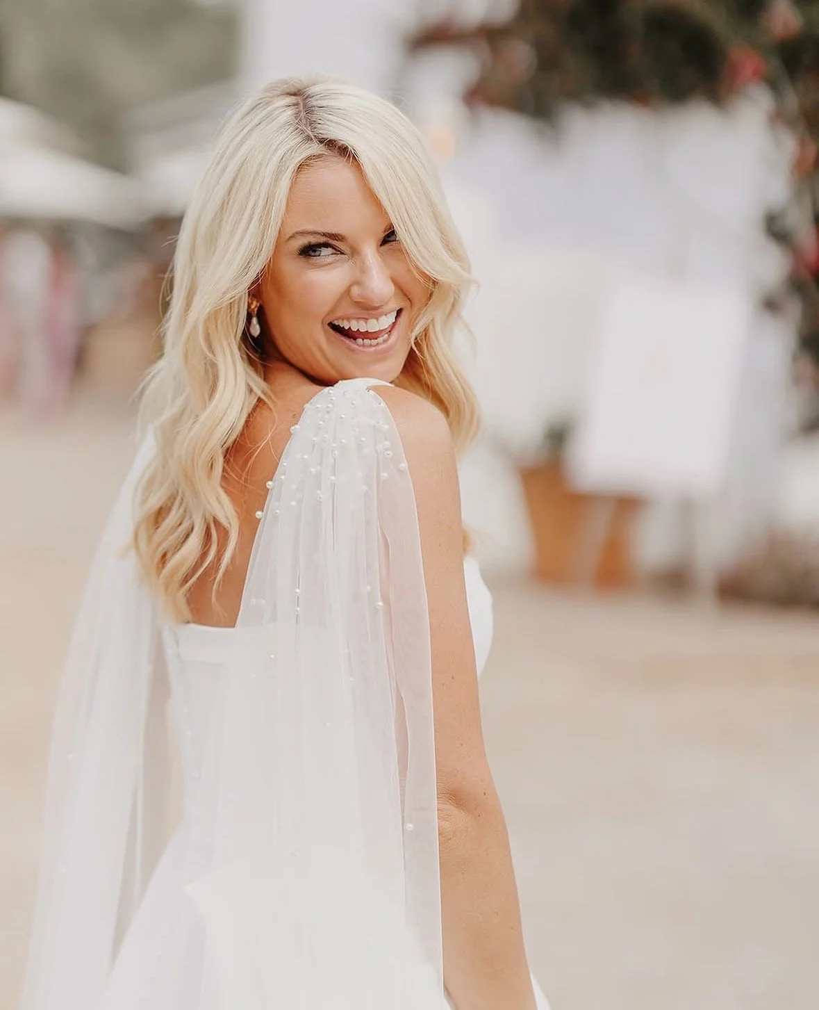 A smiling woman with long blonde hair wearing a white dress with pearl details on the shoulder, standing outdoors with blurred background.
