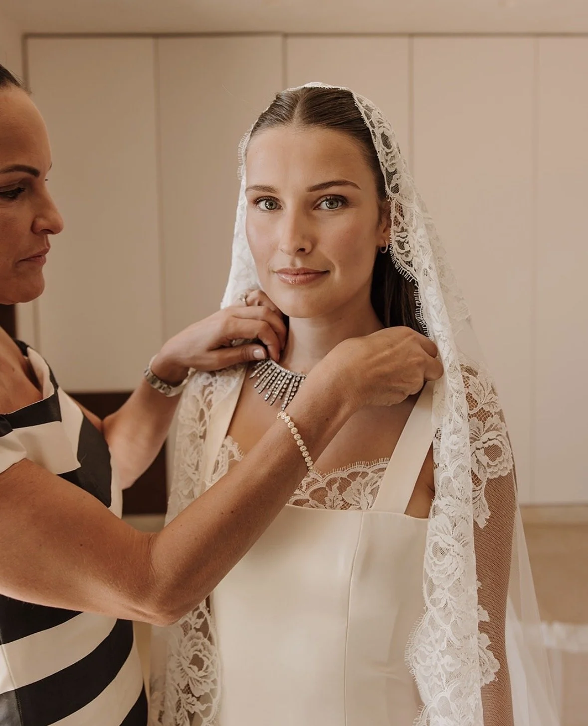 A bride with a lace veil getting ready for her wedding, assisted by an older woman adjusting her jewelry in a room.