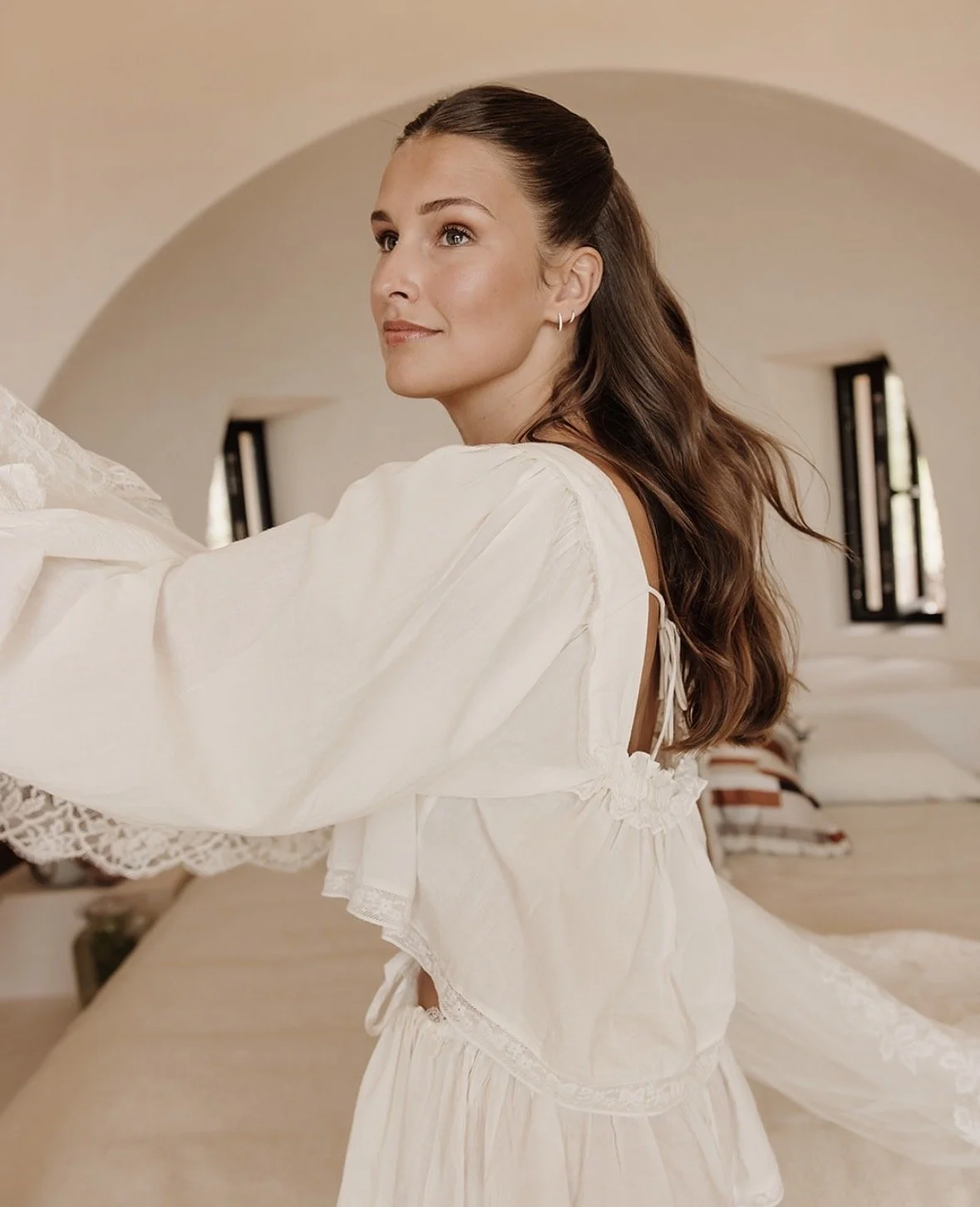 A woman with long brown hair in a half-up style, wearing a white dress, sitting on a bed in a room with an arched ceiling, looking to the side with a gentle smile.
