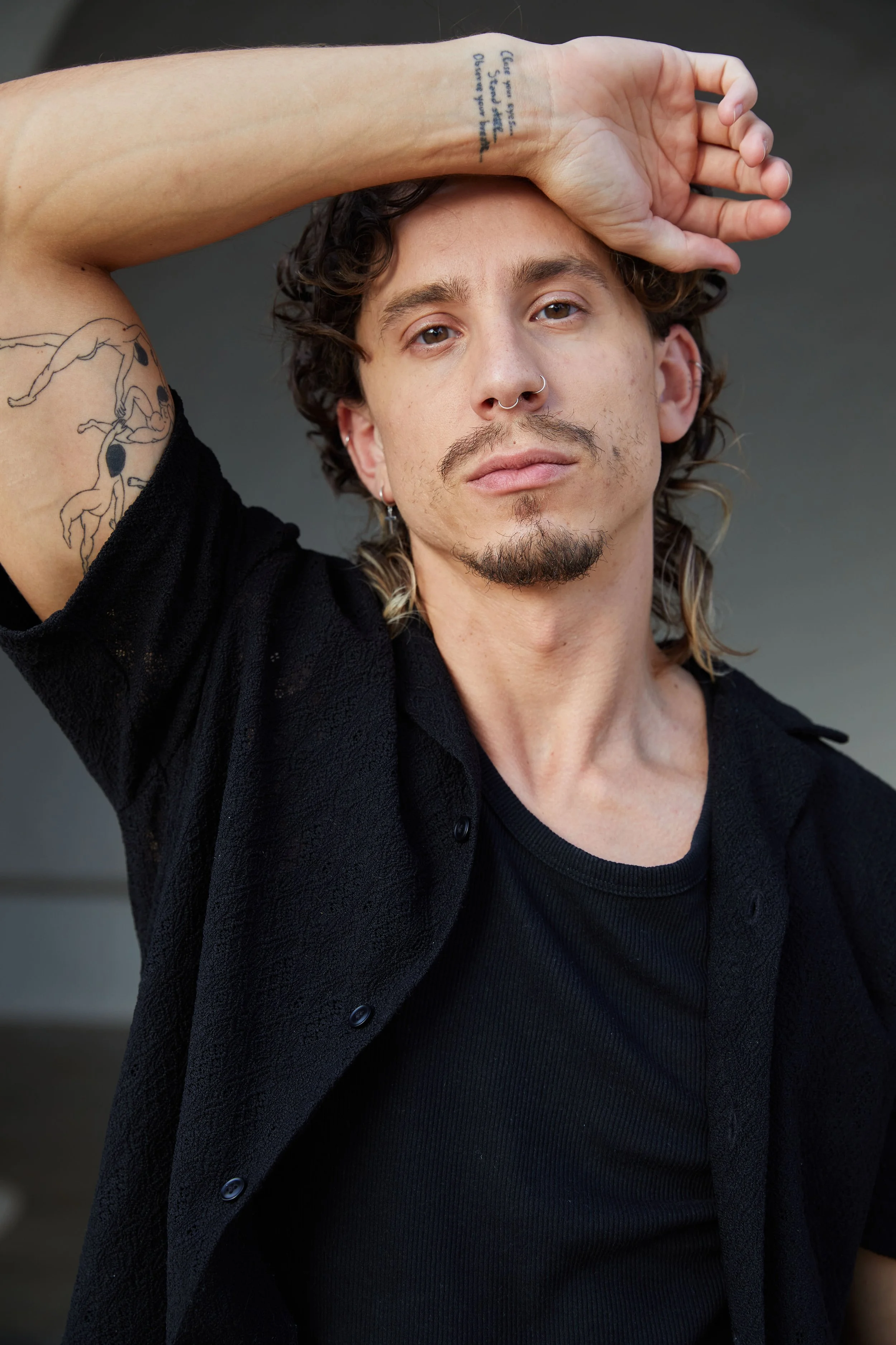 A young man with curly hair, tattoos on his arm and shoulder, and facial piercings, looking into the camera with a serious expression, wearing a black shirt with a textured black jacket over it.