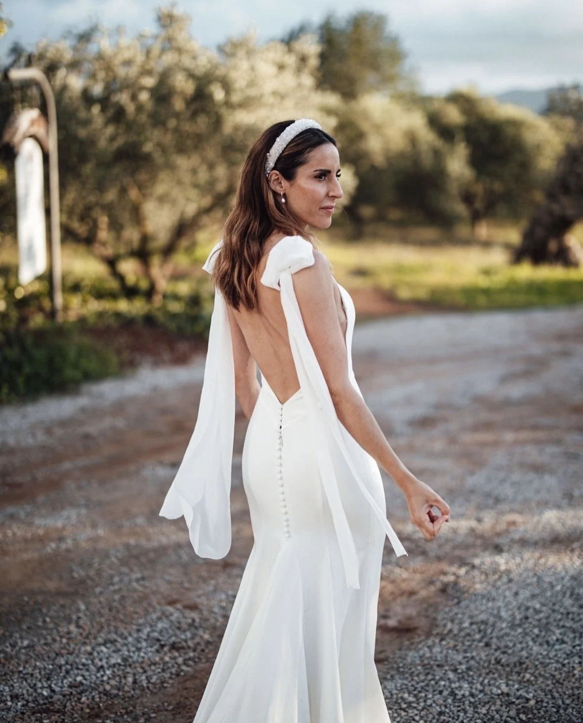 Woman in a white wedding dress with open back, walking outdoors on a dirt path, with trees and a blue sky in the background.