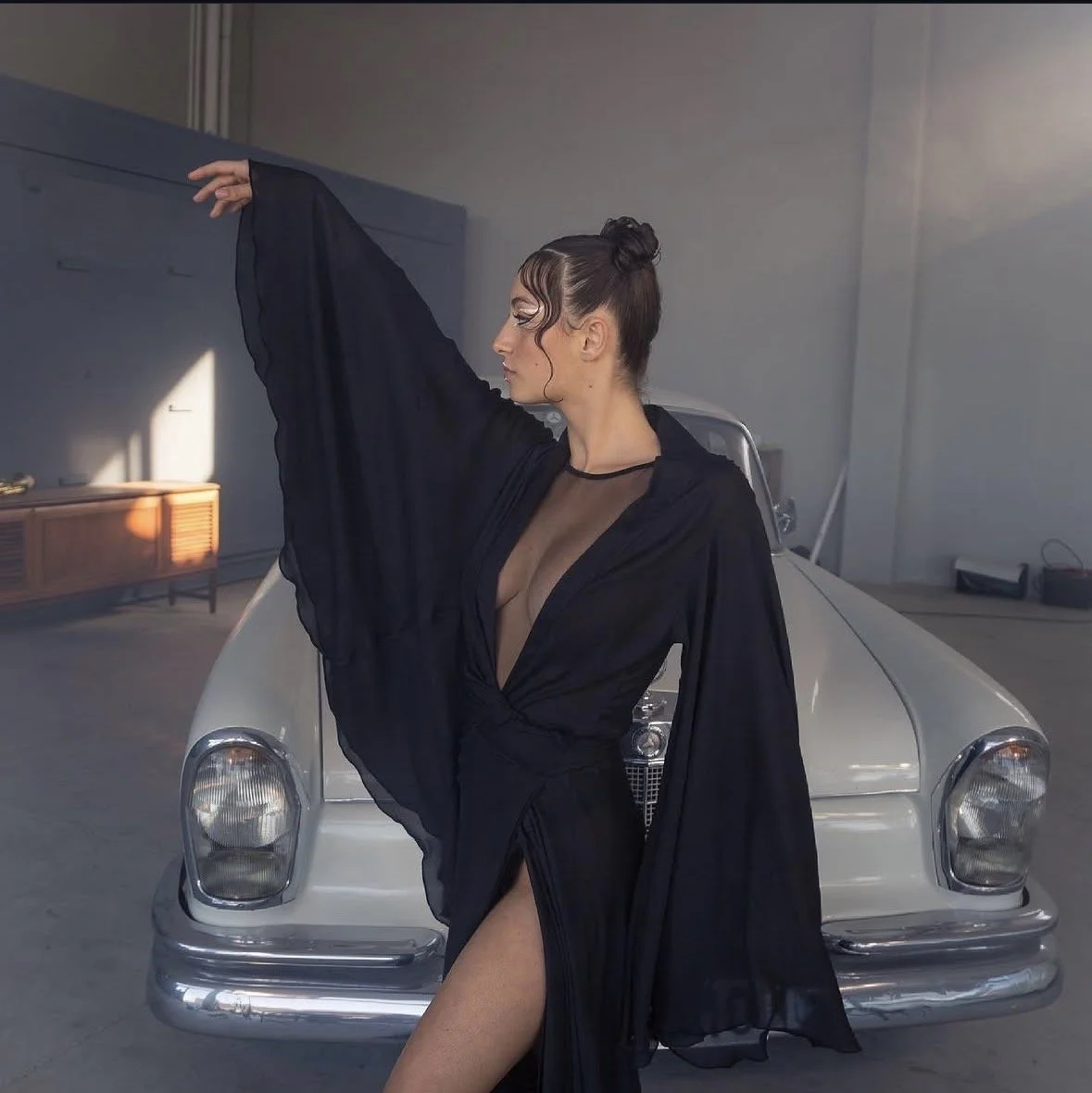 Woman with dark hair styled in an elegant updo, wearing a sheer black gown with a plunging neckline and thigh-high slit, posing in front of a vintage silver car in a spacious, minimalist garage.