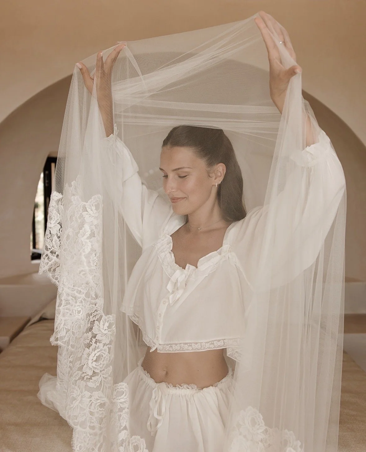 A woman with dark hair, wearing a white lacy crop top and matching shorts, holding a sheer veil above her head in a softly lit room with beige walls.