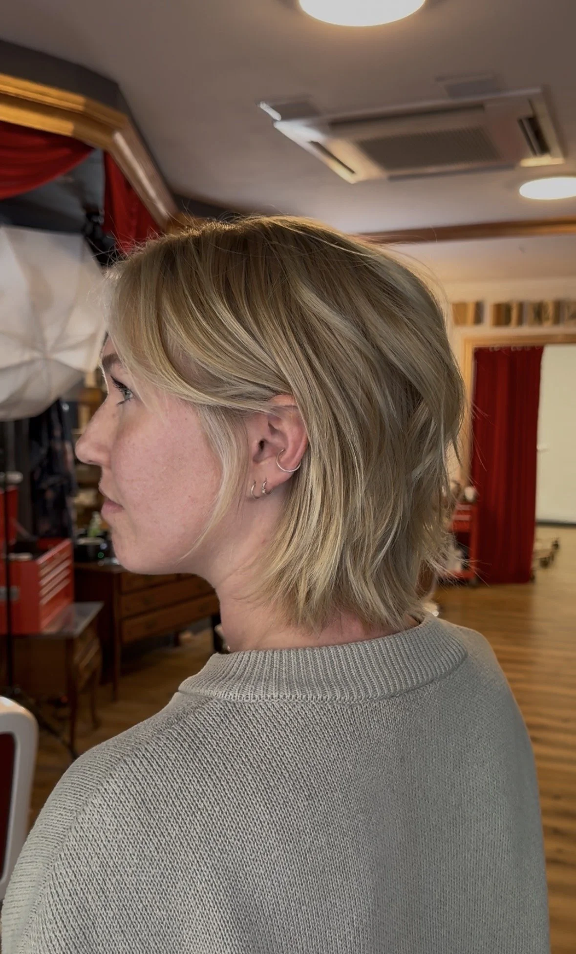 Side view of a woman with short blonde hair and earrings, standing indoors with wooden flooring, red curtains, and equipment in the background.