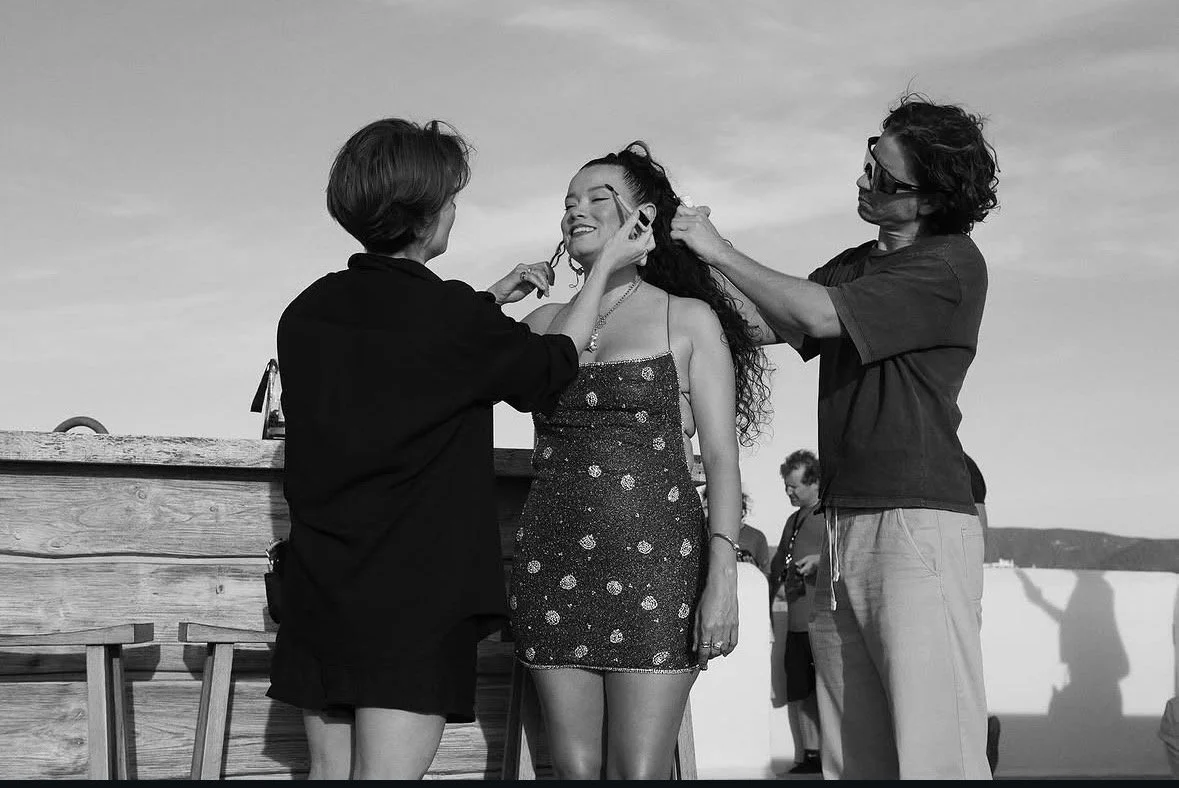 People applying makeup or styling hair to a smiling young woman outdoors, with a wooden barrier and other people in the background.
