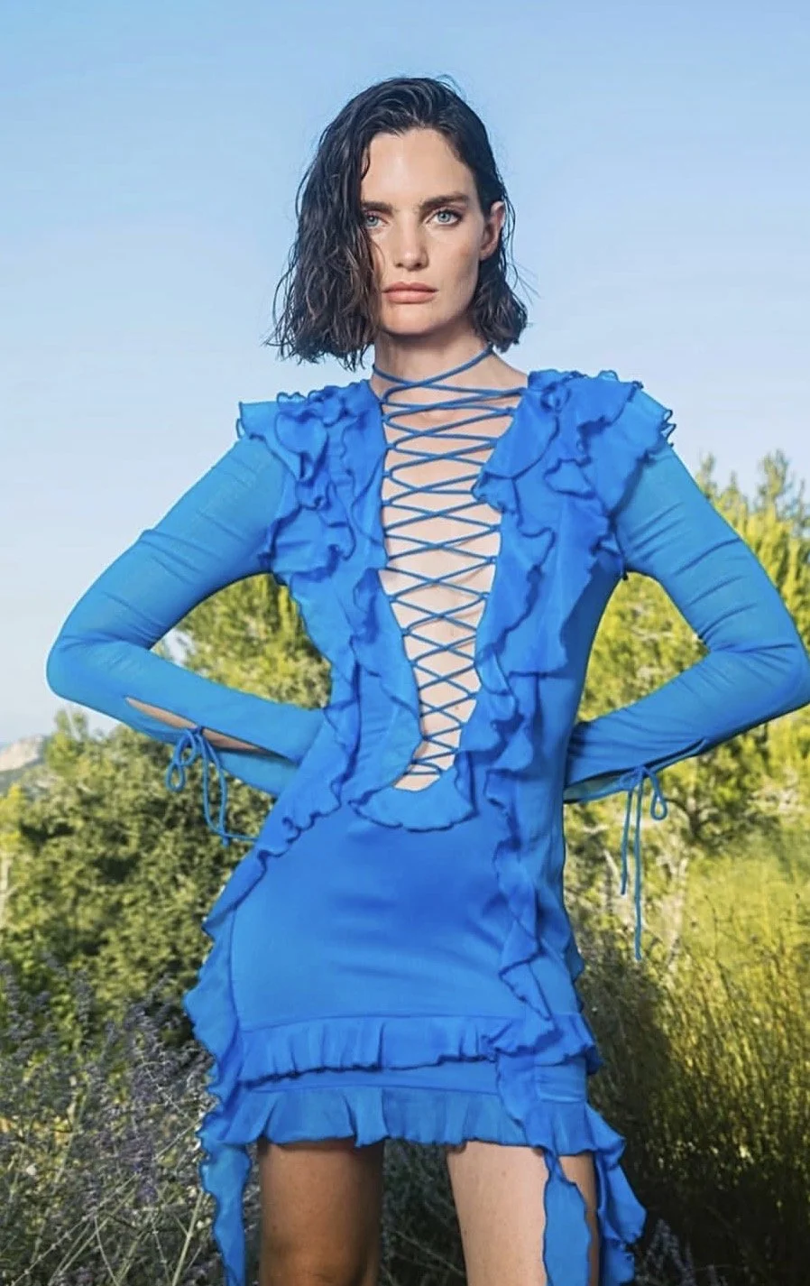 A woman with dark, wavy, shoulder-length hair posing outdoors in a bright blue dress with ruffles and lace-up details in the front. She stands with hands on her hips against a background of greenery and a clear blue sky.
