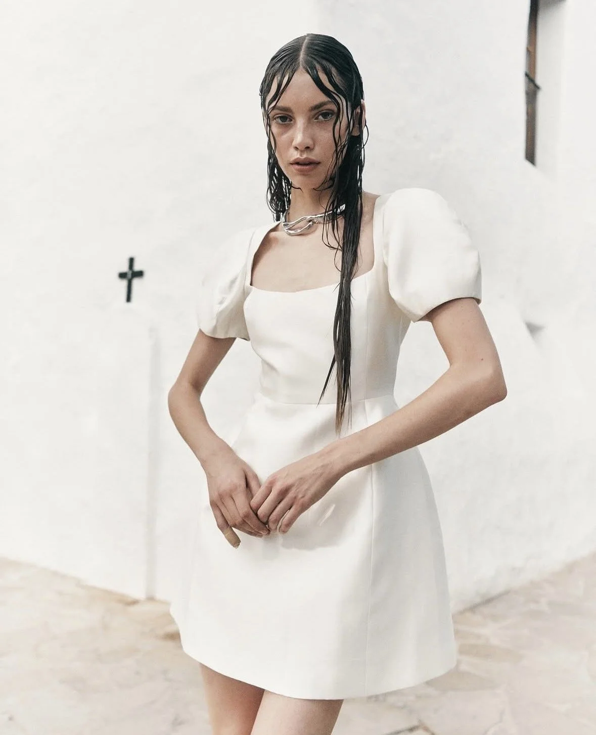 A woman with long wet dark hair wears a white dress with puffed sleeves and a chunky silver necklace, standing indoors against a white wall with a small black cross and a window.
