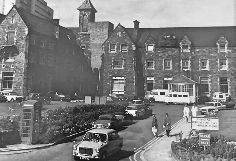 Black and white photo medical facility with multiple cars parked outside, a telephone booth, and a sign that reads 'Parking for the Samaritan Medical Staff or Ambulance Only.' Several people are walking near the entrance, and a bus is parked on the side.