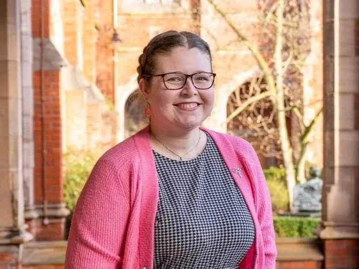 Dr. Robyn Atcheson in front of the Lanyon Building at Queen's University Belfast, wearing glasses, a pink cardigan, and a black and white patterned dress.