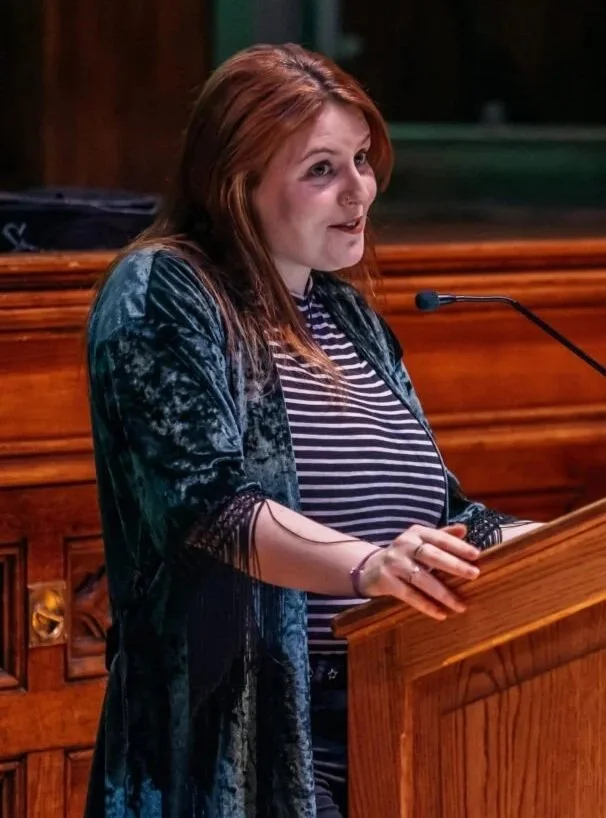 Dr. Rebecca Watterson delivering a talk in the Guildhall, Derry. Dr. Watterson has red hair is speaking at a wooden podium with a microphone, in a room with wooden paneling.