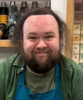 Leading historian of the second world war in Northern Ireland Michael Burns. A bearded man with curly hair smiling, wearing a green shirt, in a workshop with shelves of bottles and boxes in the background.