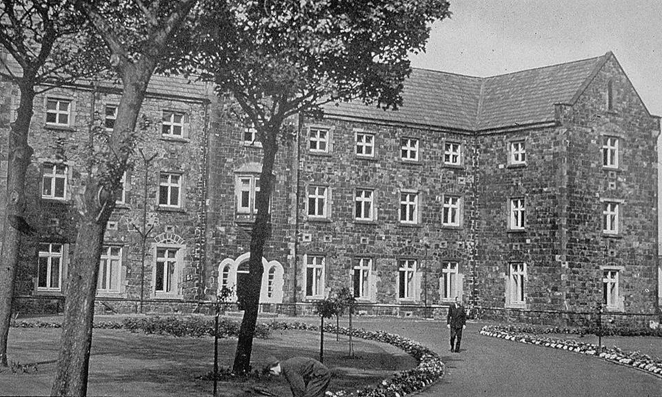 A view of the Belfast Union Workhouse fever hospital. The photograph is black and white with trees in the foreground, behind these an imposing stone built 3 story structure. 