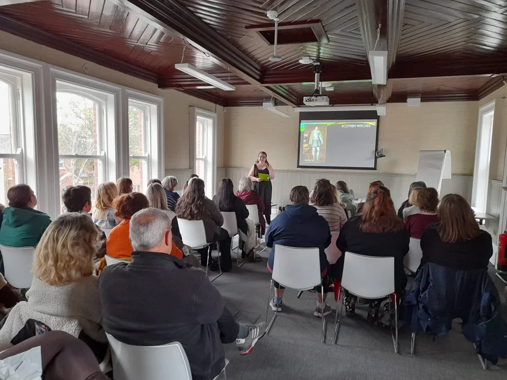 Dr. Robyn Atcheson is giving a presentation to an audience in a room with large windows, a wooden ceiling, and a projector screen displaying a slide titled 'Scottish Witches' with an image of a historical figure.
