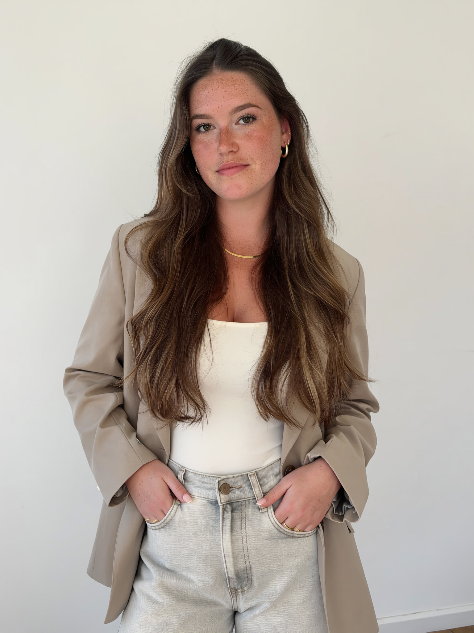 Social Media Manager, content creator. Portret van een jonge vrouw met lang bruin haar en sproeten, gekleed in een beige blazer, witte top en lichtgekleurde spijkerbroek, staand voor een witte achtergrond.