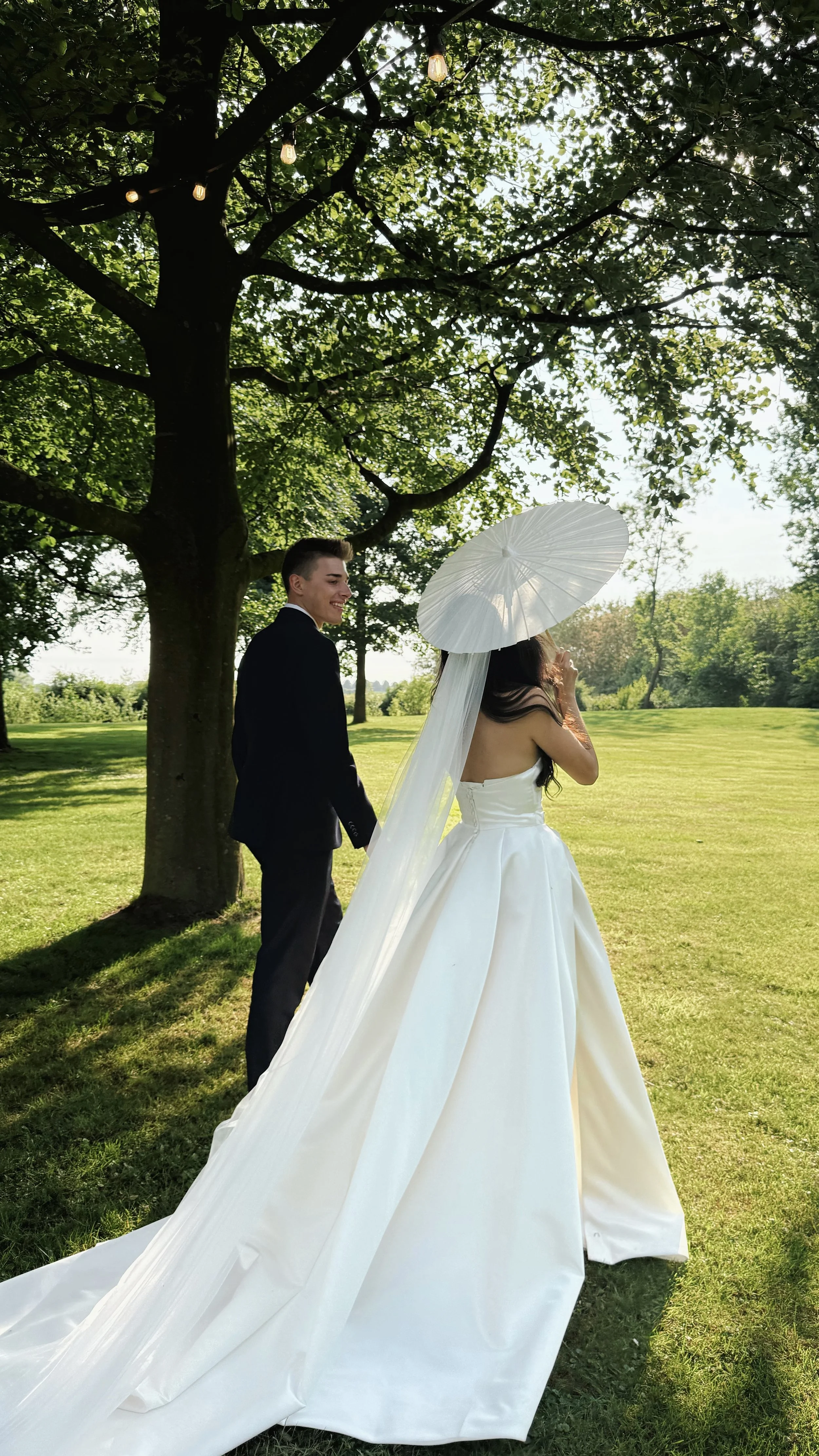 Wedding Content Creatie Vlaanderen, België. Een bruid in een witte jurk en een bruidegom in een zwarte pak staan buiten onder een grote boom met groene bladeren, met een witte parasol en zonne-energie in een park of tuin.