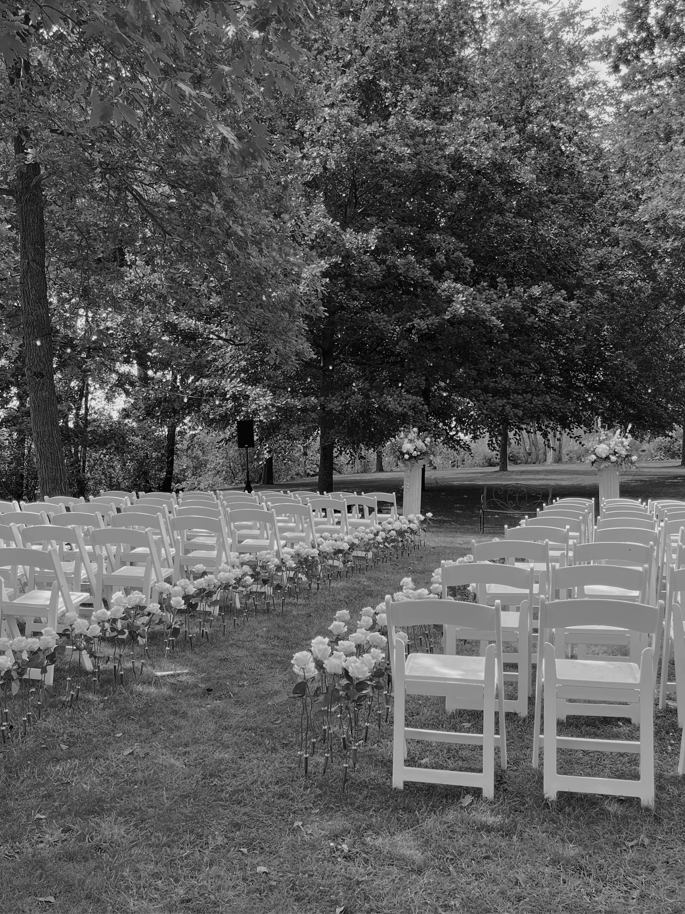 Wedding Content Creatie Vlaanderen, België. Een buiten ceremonie setup met witte stoelen en bloemendecoraties onder grote bomen in een park of tuin.