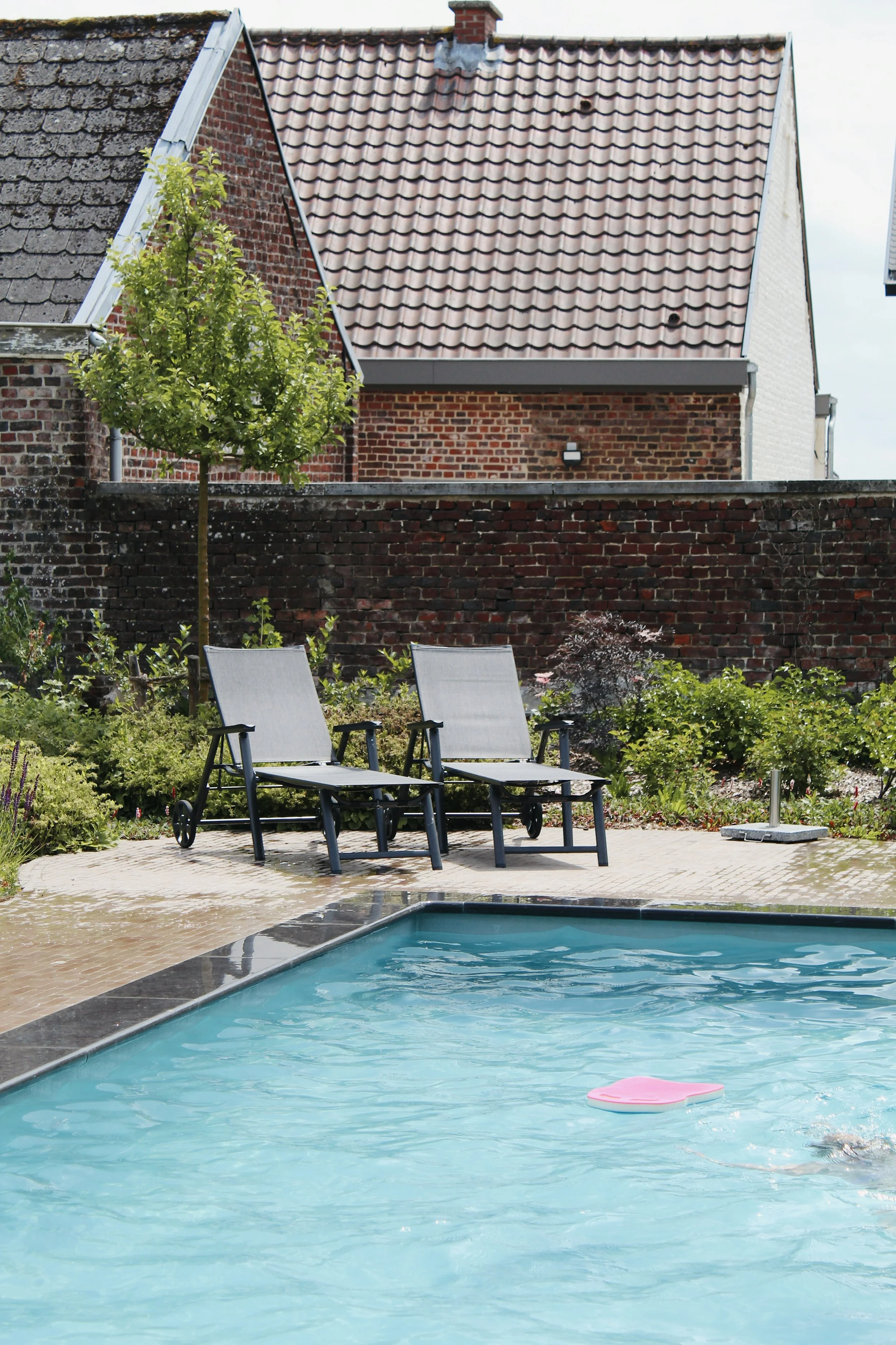 Een zwembad met een ligstoel en een opblaasbare zwemband op het water, omringd door een betegeld terras en een tuin met planten en een boom, met een bakstenen huis met een rood pannendak op de achtergrond.