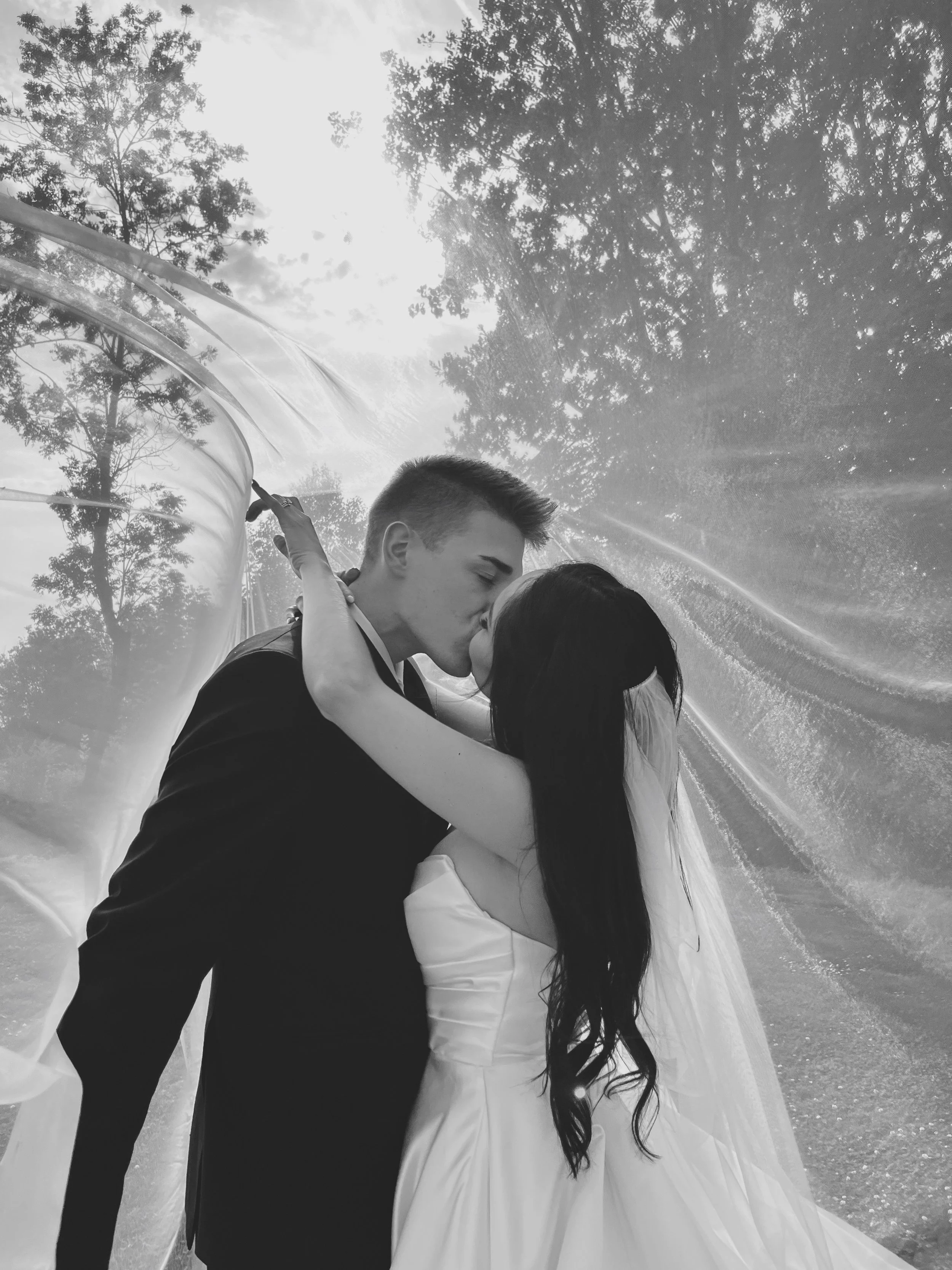 Wedding Content Creatie Vlaanderen, België. Een bruid en bruidegom kussen elkaar buiten, omringd door bomen en natuurlijke lichtstralen.