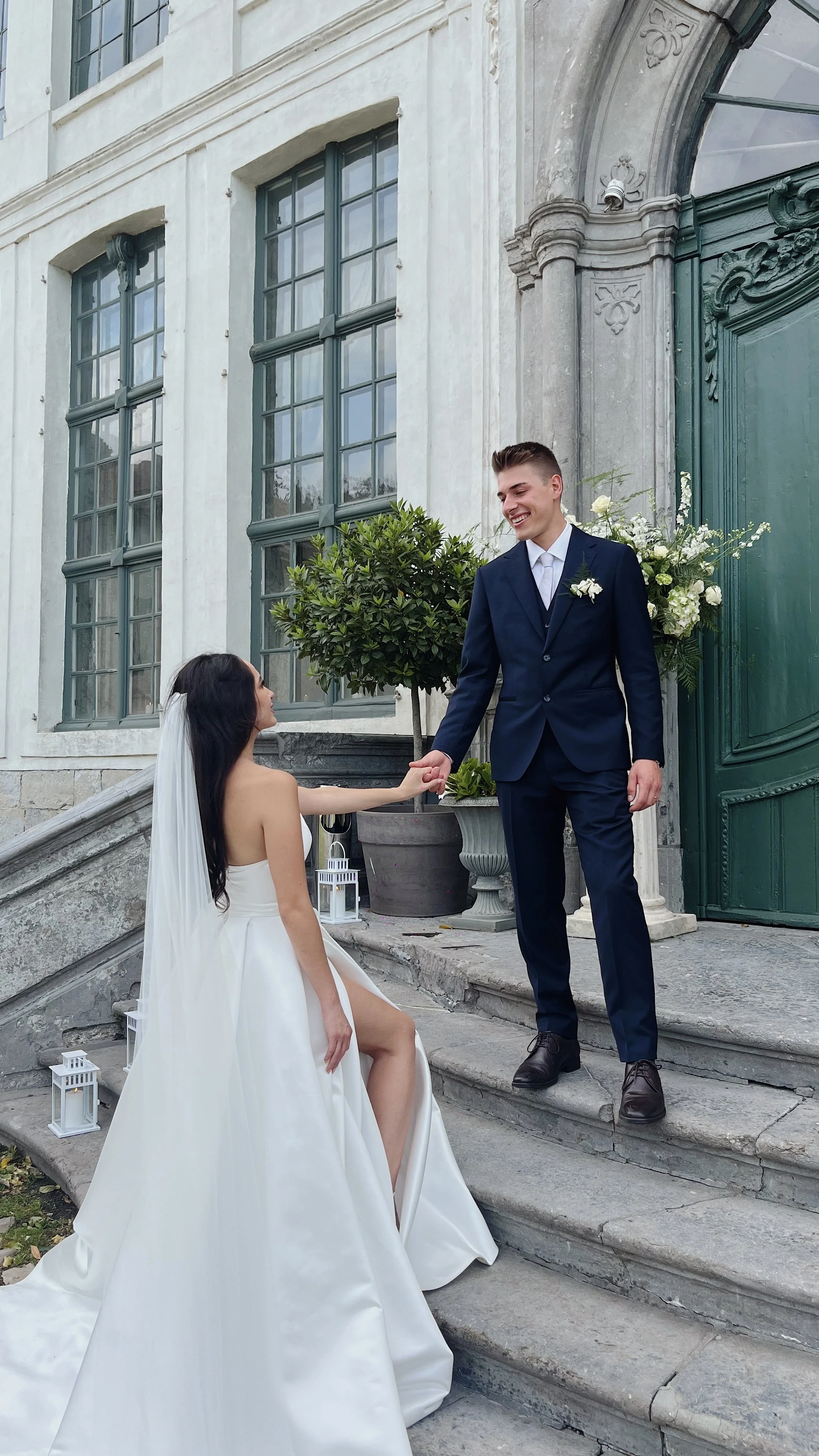 Wedding Content Creatie Vlaanderen, België. Een getrouwde koppel uitwisselt huwelijksgeloften op de trap voor een historisch gebouw. De vrouw zit in een witte trouwjurk en de man in een donkerblauw pak, terwijl ze hand in hand staan en glimlachen.