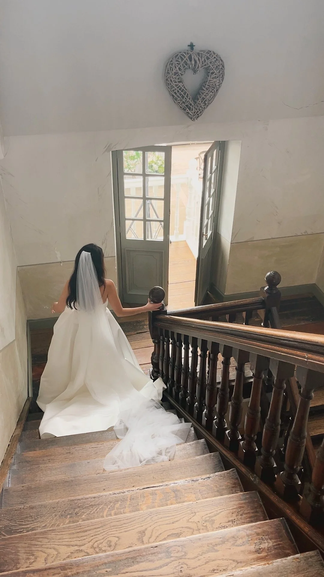 Wedding Content Creatie Vlaanderen, België. Vrouw in een witte trouwjurk met lange trein staat op een houten trap, haar hand rust op de nieuweel bruine balustrade, en kijkt naar buiten door een openstaande deur.