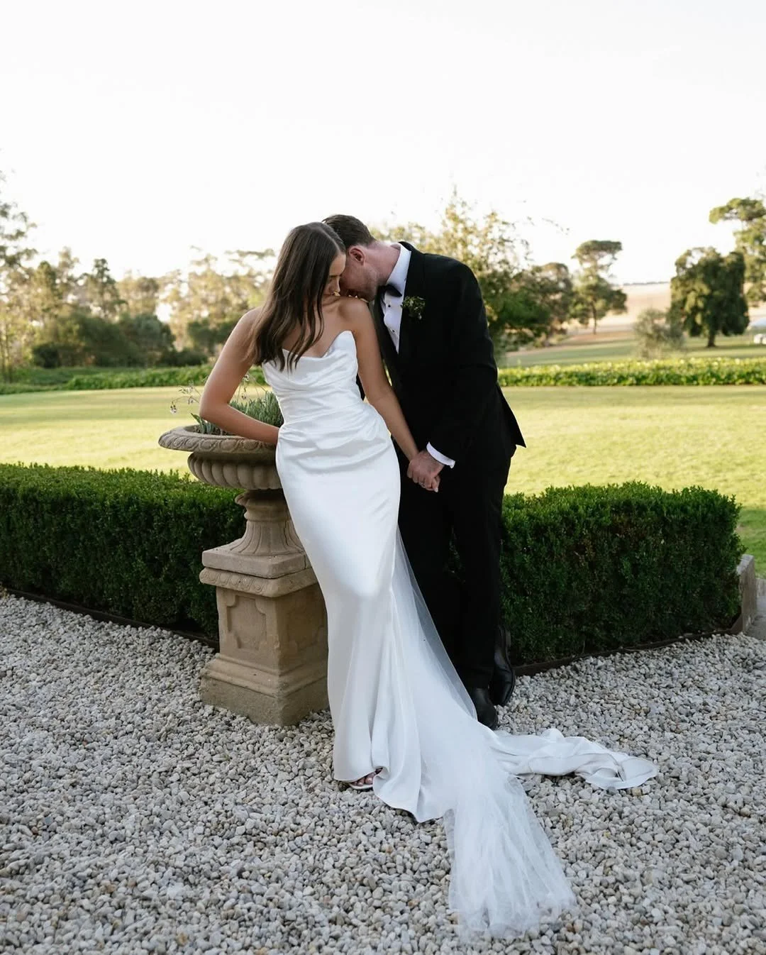 Wedding Content Creatie Vlaanderen, België. Een bruid en bruidegom op een witte stenen patio, schouder aan schouder, bij een tuin met groene gazons en bomen, in formele trouwkleding, tijdens een fotoshoot.
