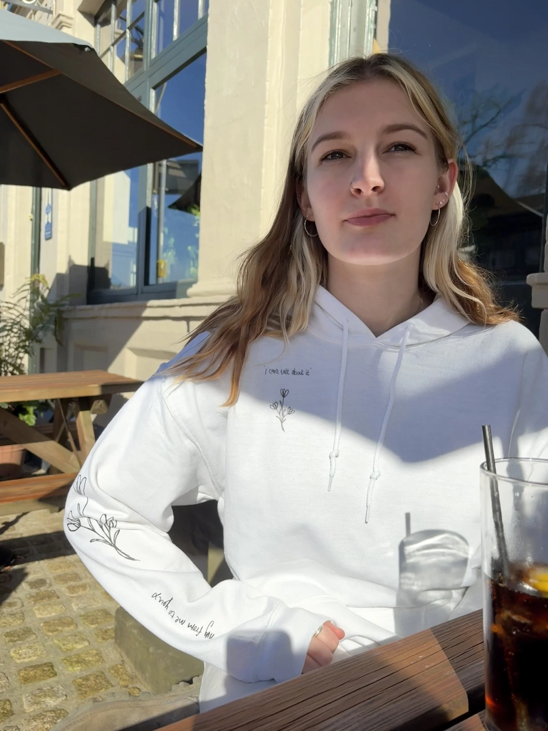 A young woman with blonde/brown hair, wearing a white hoodie with small flower and text designs, sitting outdoors at a wooden table with her drink in front of her, in a bright, sunny setting.