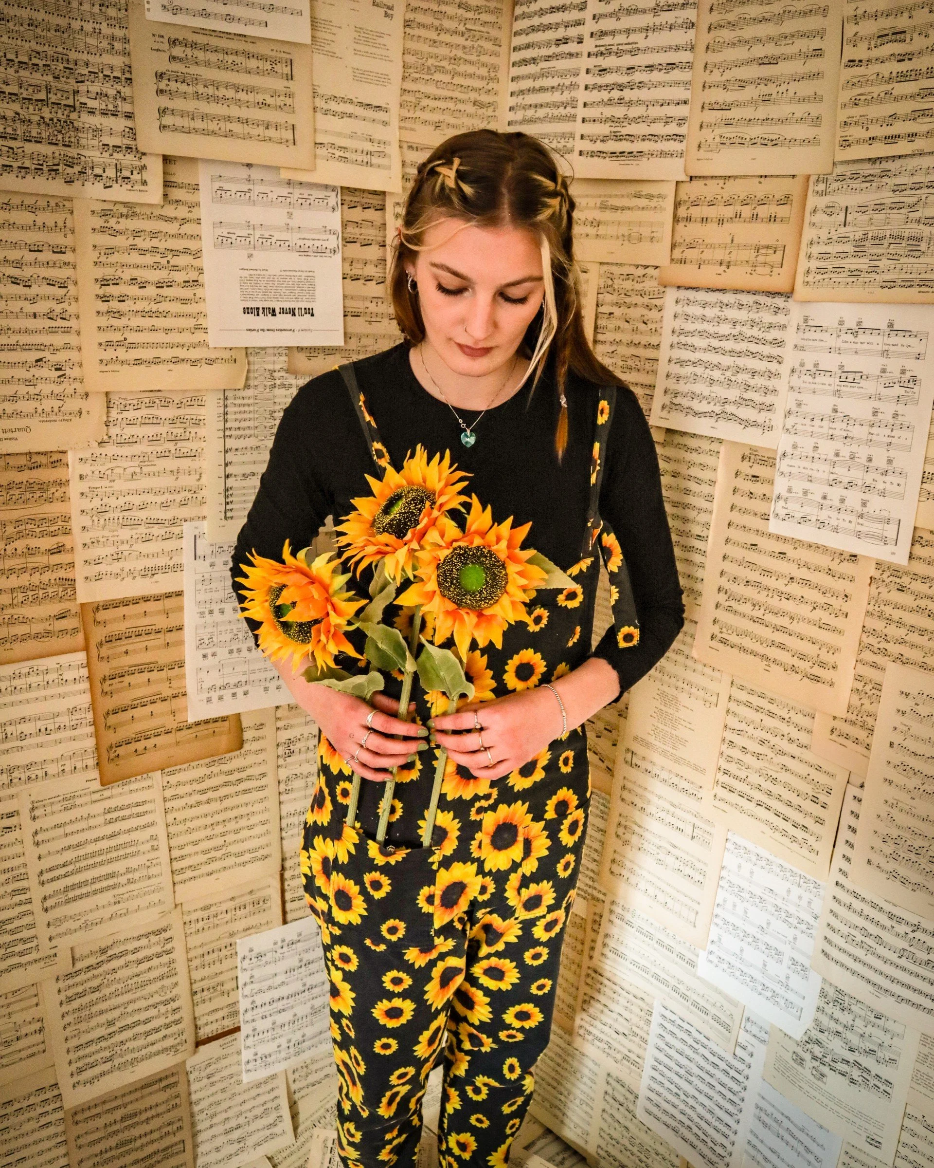 A young woman with long brown hair standing in front of a wall covered with sheet music, wearing sunflower dungerees, and holding a bouquet of sunflowers.