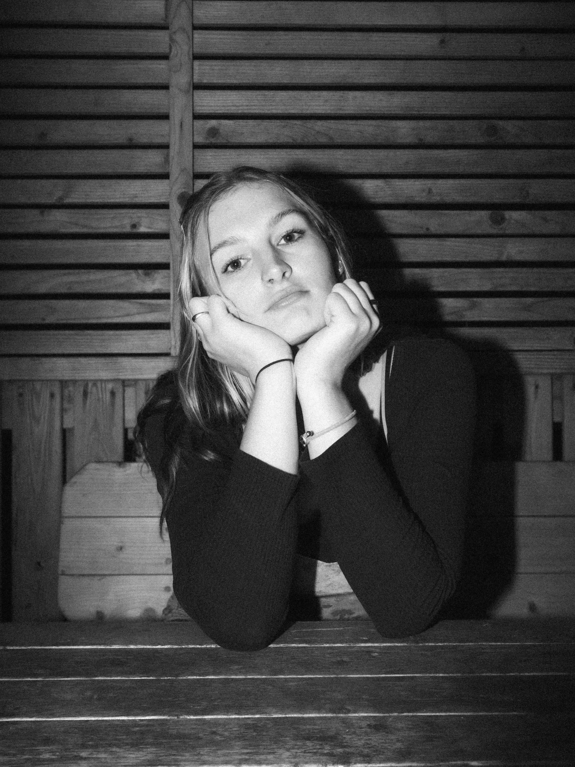 A young woman with long hair, resting her face on her hands, looking directly at the camera. She is seated at a wooden table with a wooden wall in the background.