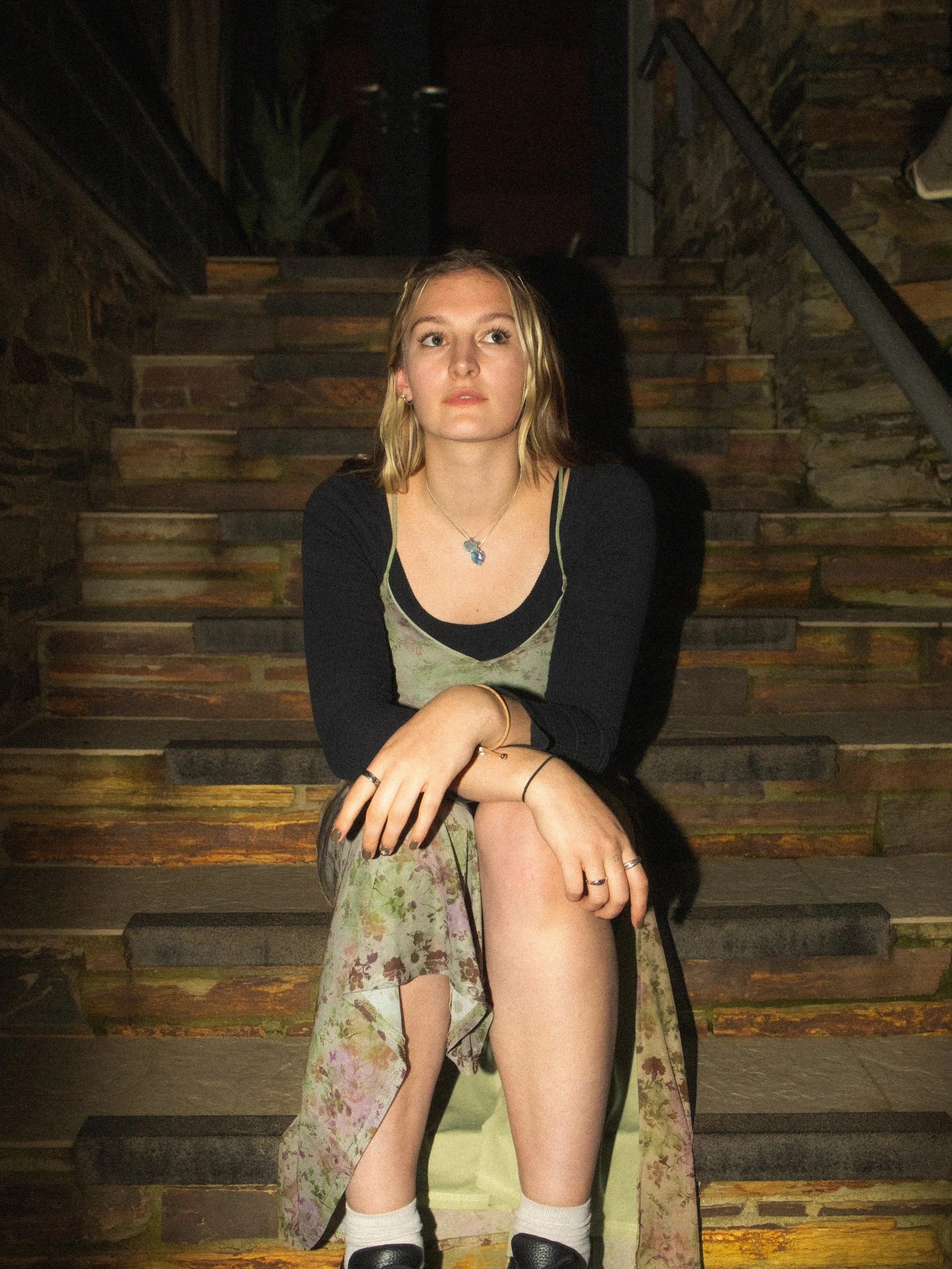 A young woman with blonde/brown hair sitting on a stone staircase, wearing a floral dress with a black top underneath, accessories, and black shoes. It is night and she's illuminated by a nearby light.